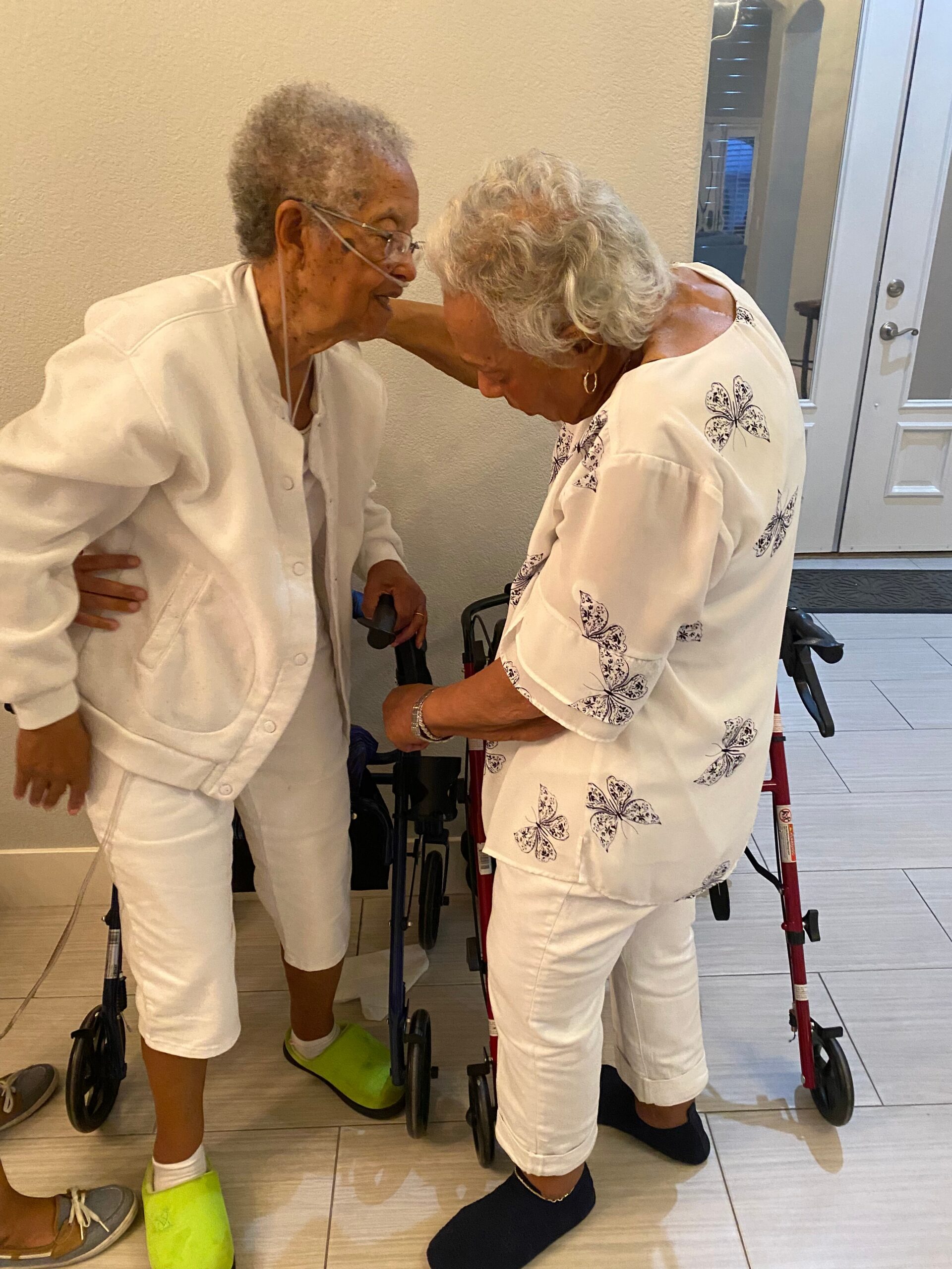 Two elderly women in white outfits share a caring moment, leaning toward each other with walkers nearby in a sunny indoor hallway.