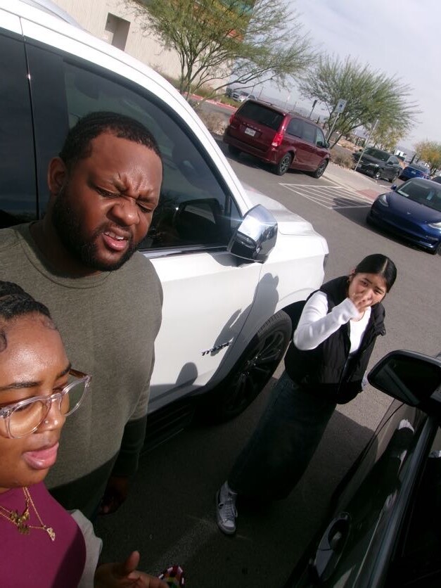 Three people in a parking lot beside a white pickup truck; one man squints, a woman with glasses smiles, another woman covers her mouth.