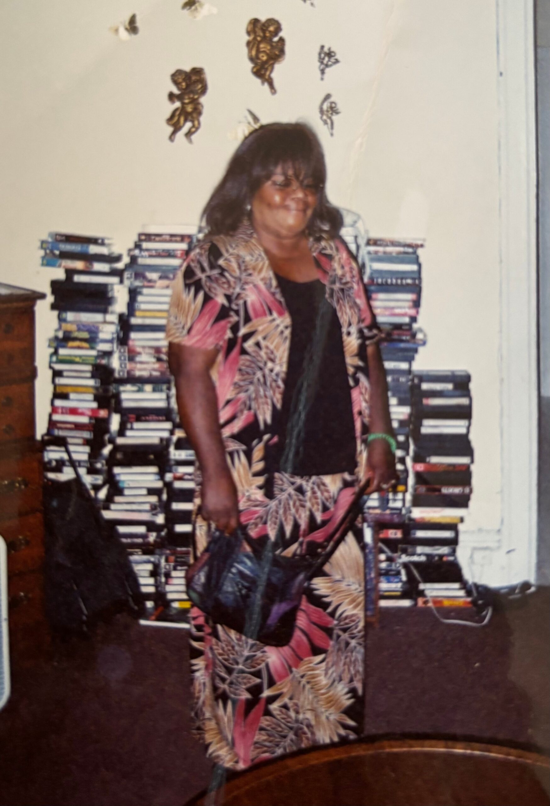 Smiling woman in a vibrant tropical-print outfit stands in front of tall stacks of CDs in a living room with butterfly wall decor behind her.