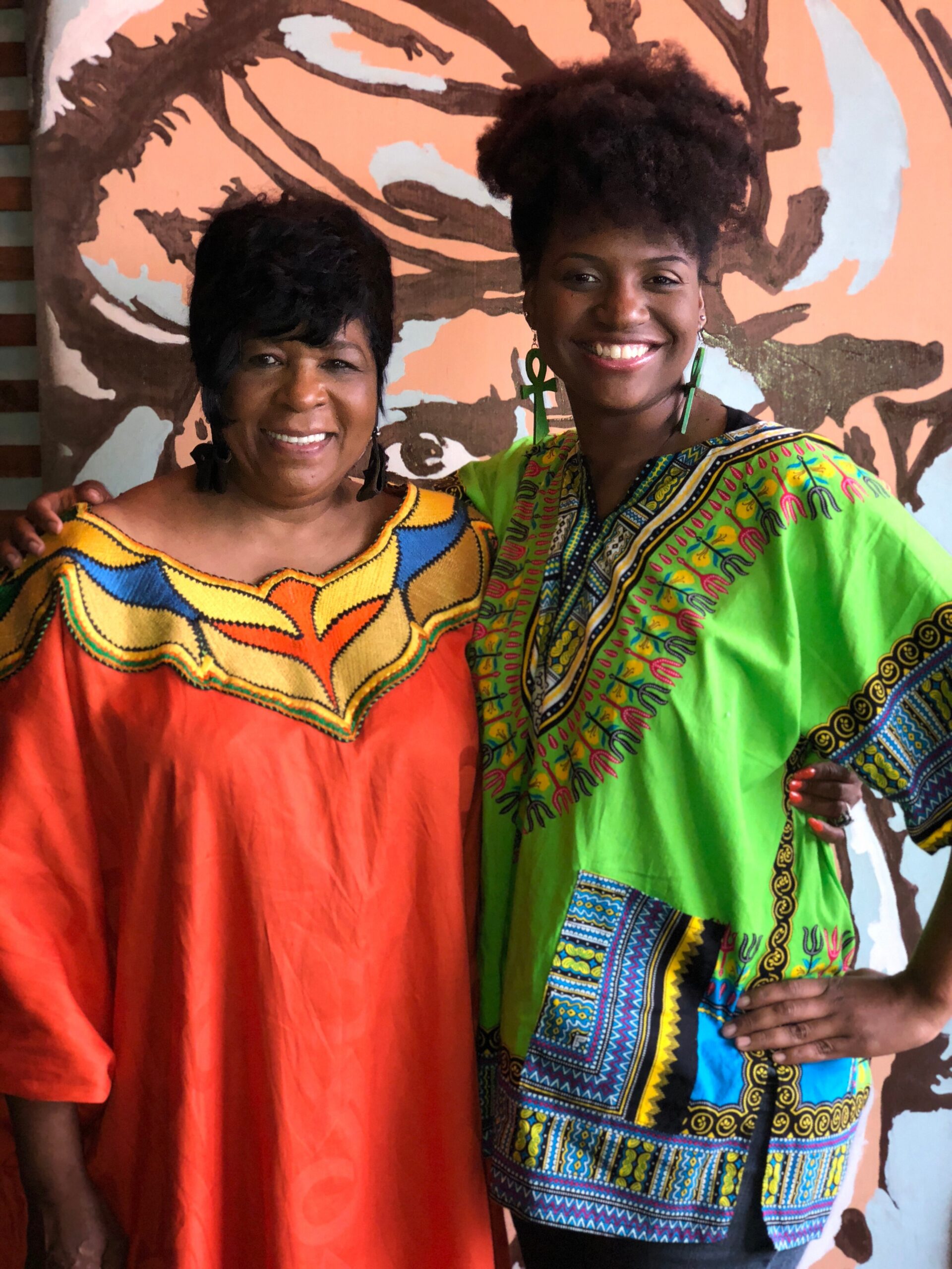 Two women posing together with happy smiles; one in an orange dress and the other in a bright, patterned dashiki, arms around each other in front of a mural backdrop.