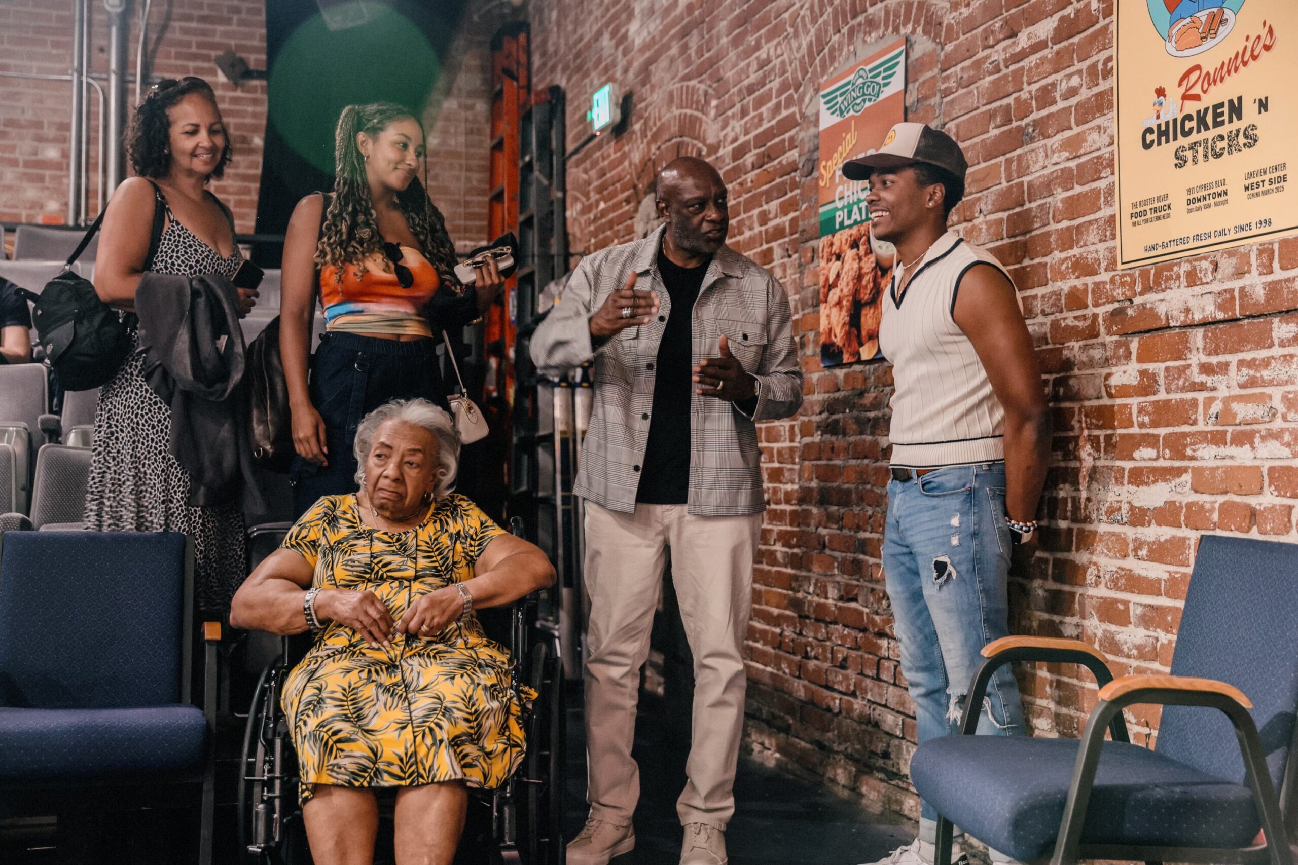 Elderly woman in a yellow tropical dress sits in a wheelchair in the foreground while four friends chat near a brick wall and blue chairs behind her.