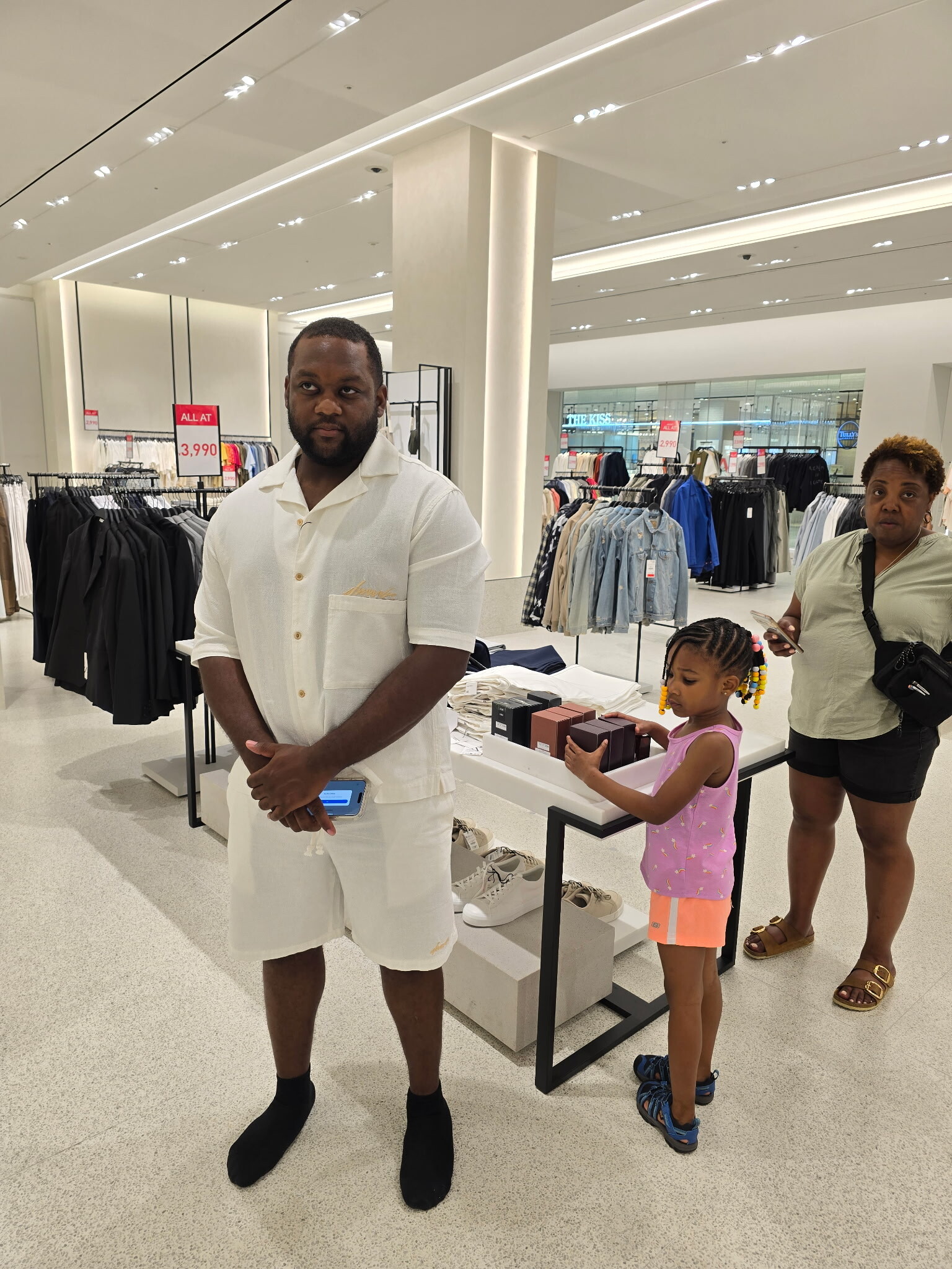 Man in light beige short-sleeve shirt and shorts stands in a bright clothing store with racks of apparel behind him; a woman and child browse at a display nearby.