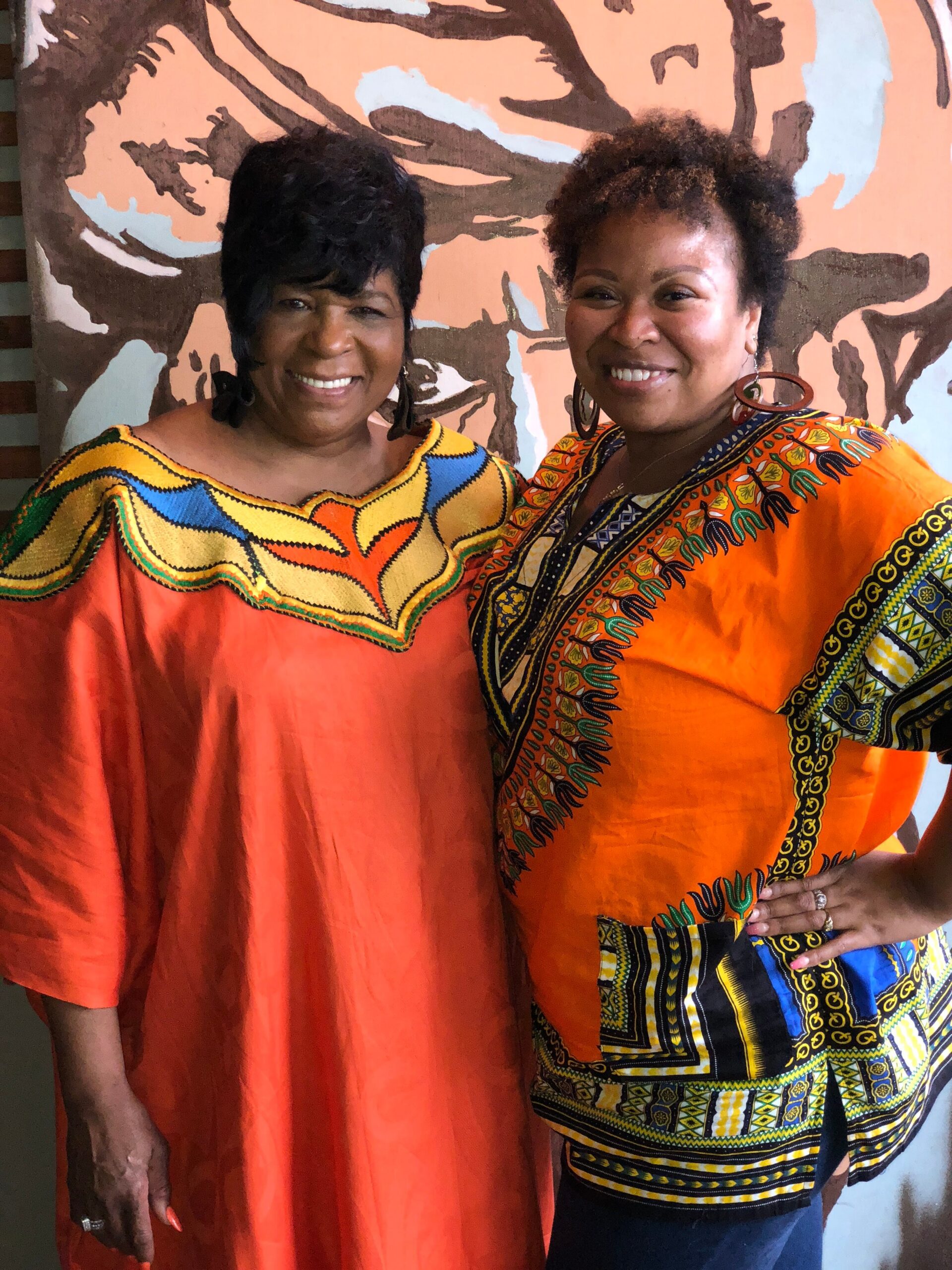 Two women smiling and posing together in bright African-print outfits, with a pink mural behind them.