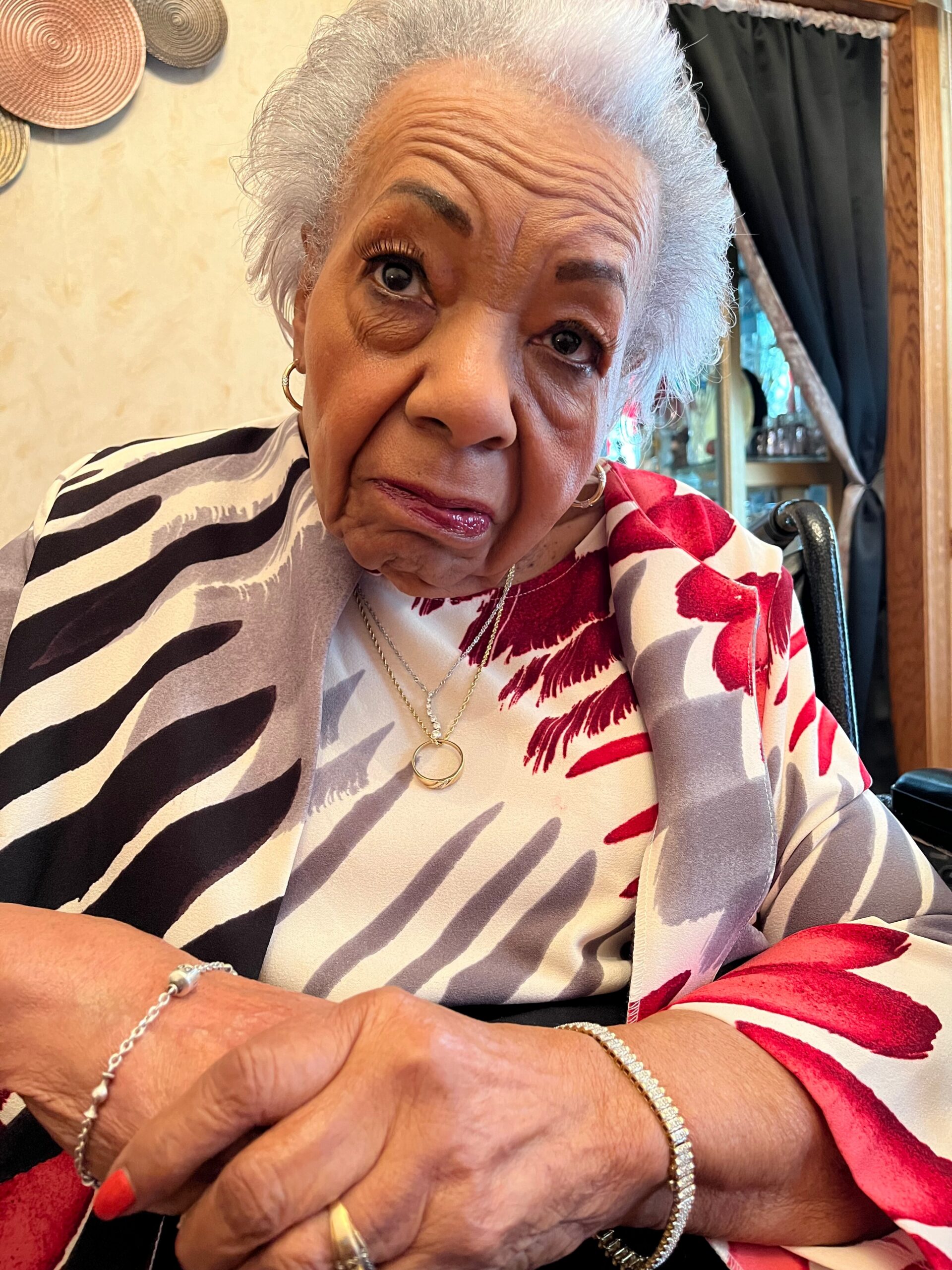 Close-up of an elderly woman with short gray hair, wearing a patterned blouse and gold jewelry, resting her arms on a table.
