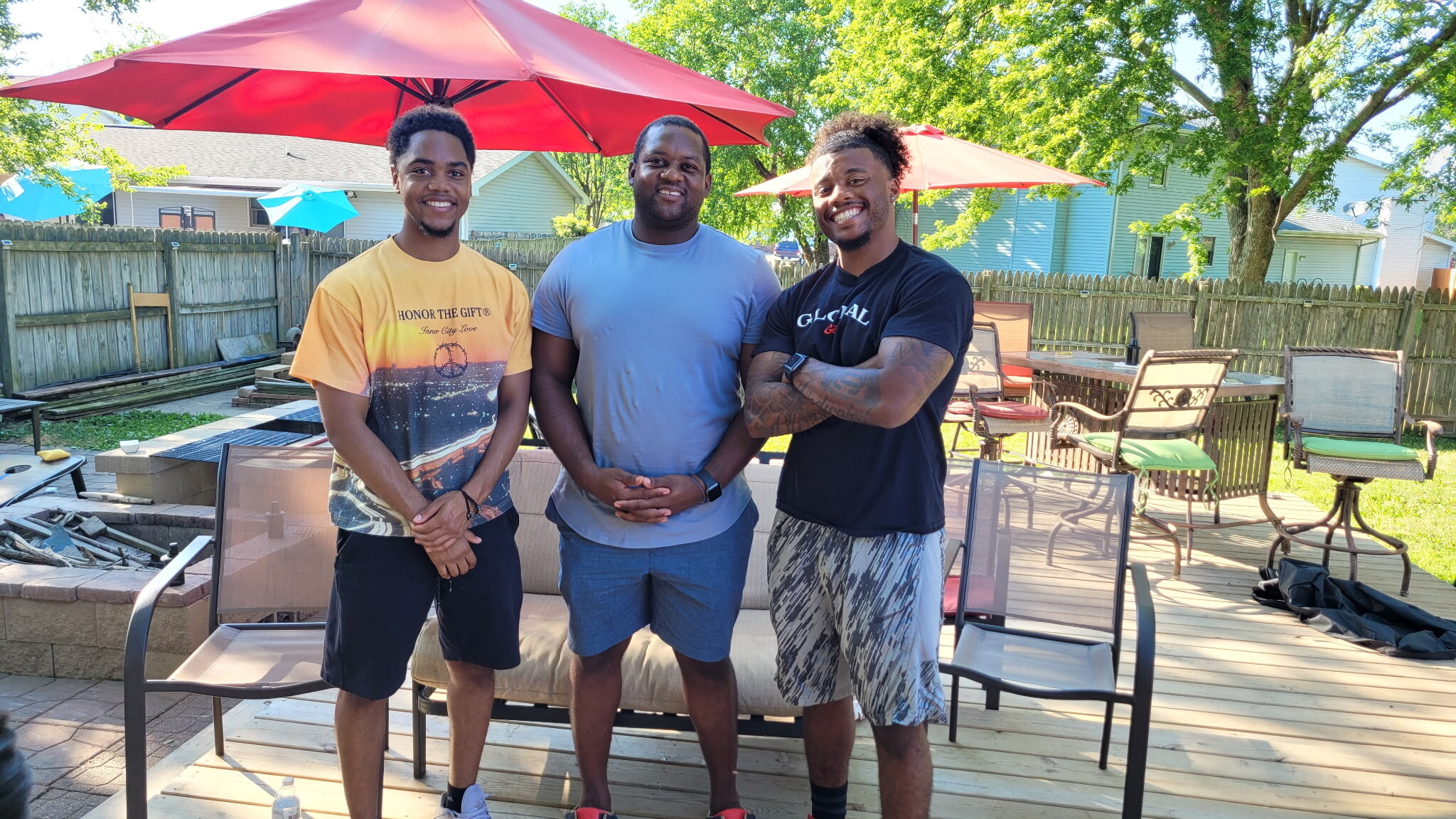 Three men pose together in a sunny backyard, smiling under red patio umbrellas with outdoor furniture and a wooden fence behind them.