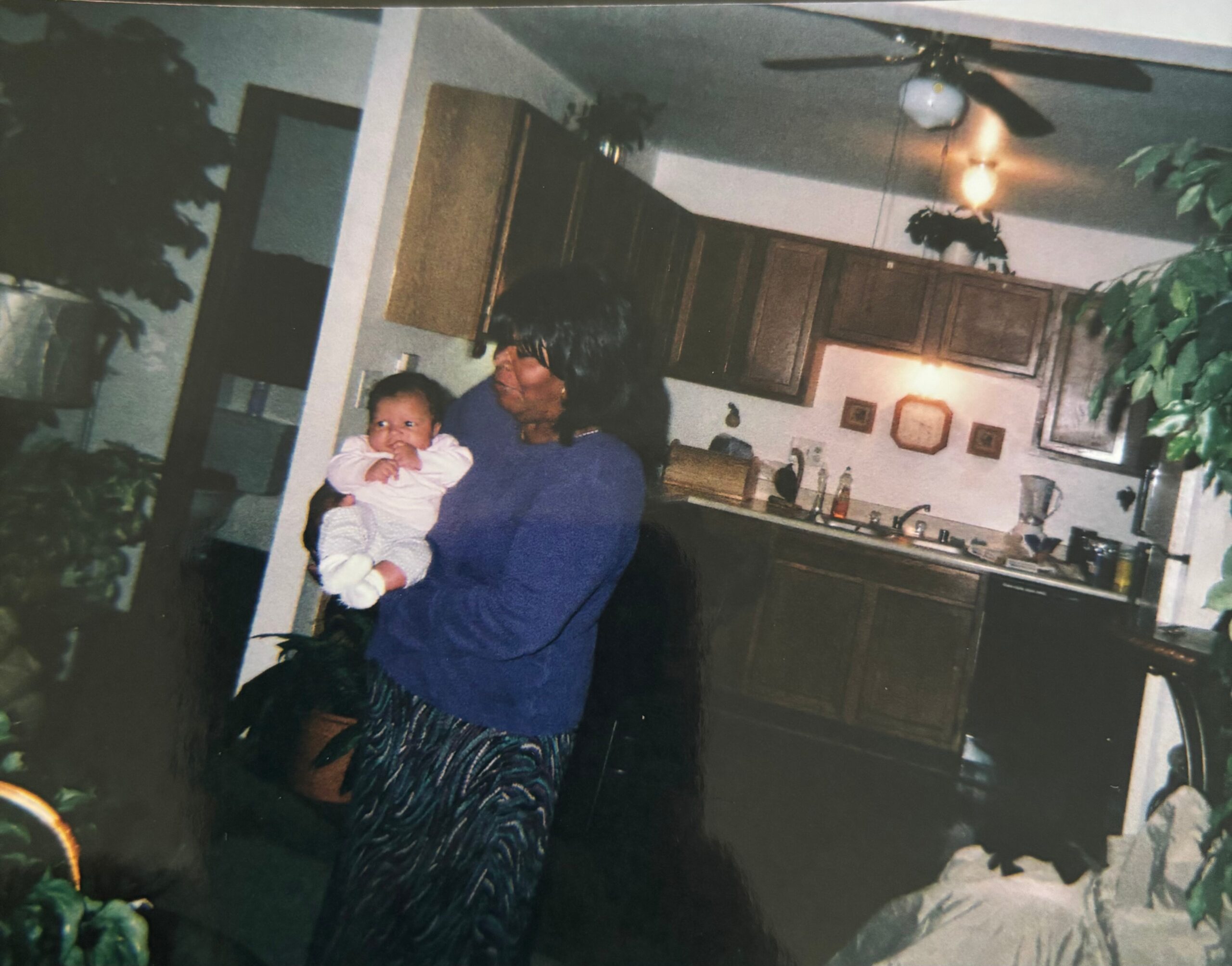 Woman in a blue top holding an infant in a home kitchen with wooden cabinets and a ceiling fan in the background.