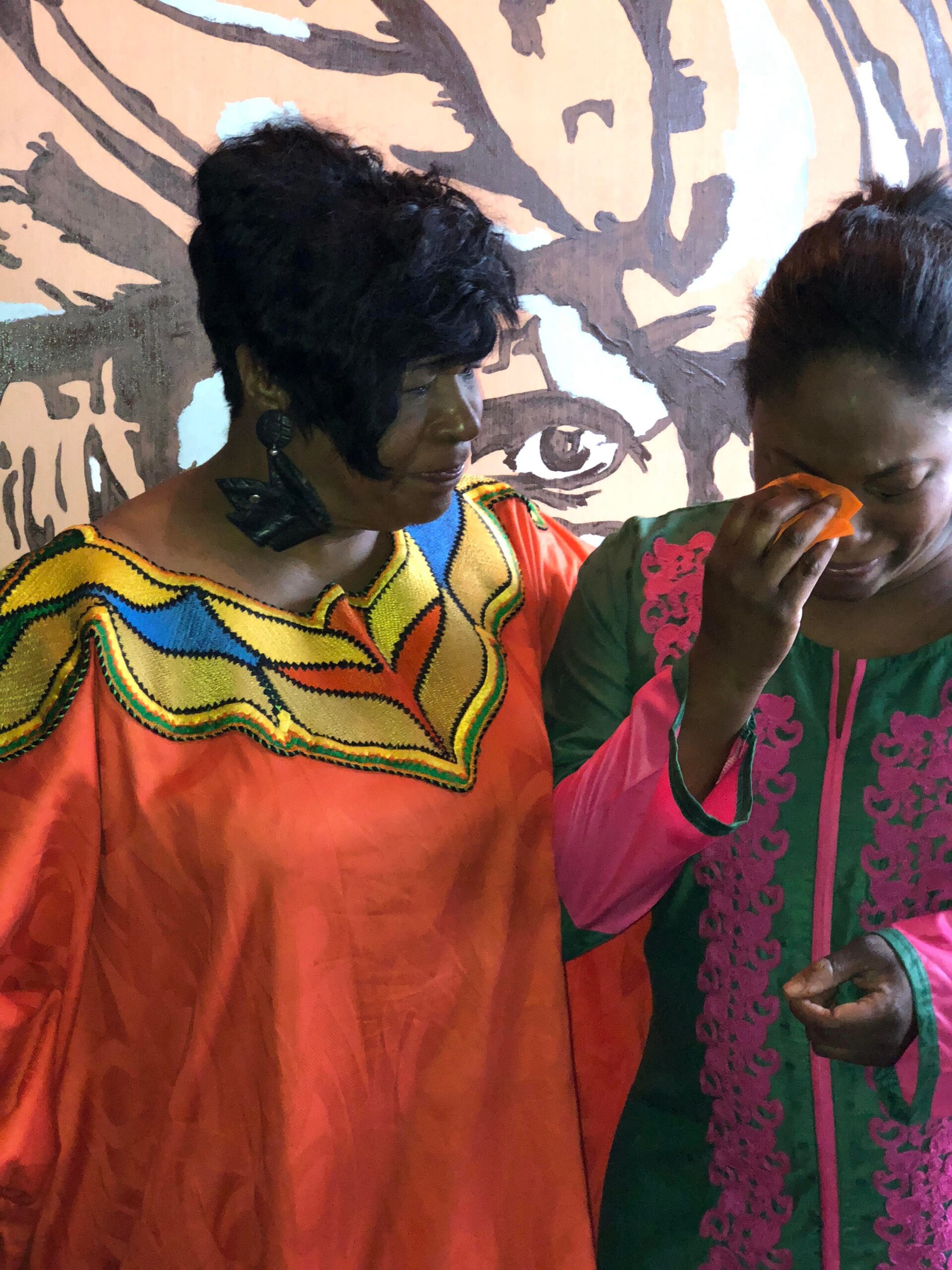 Two women in bright traditional dresses stand together, one in orange with colorful embroidery and the other in green with pink trim, in front of a mural featuring a large eye image.