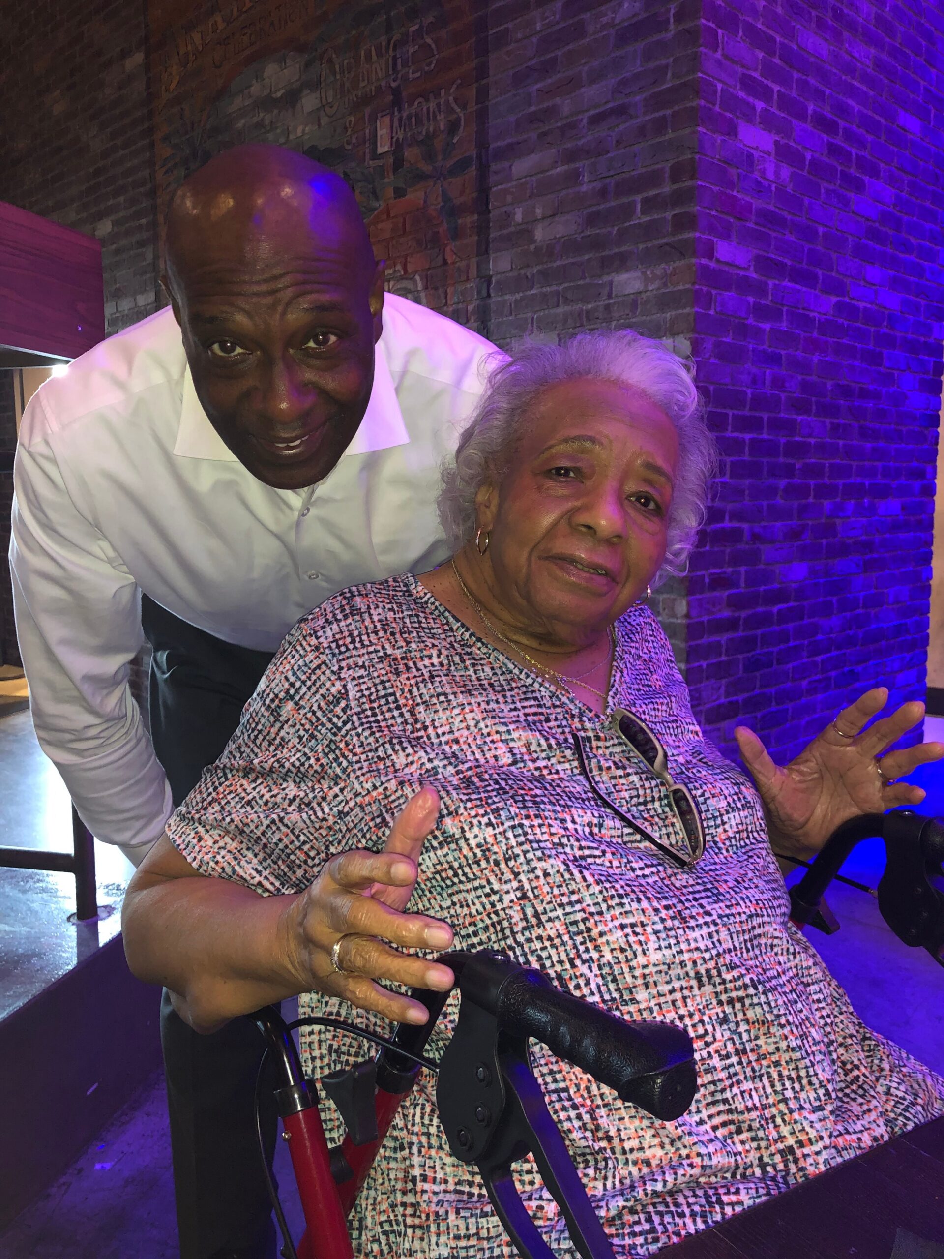 Old woman in a patterned dress seated in a wheelchair, making a peace sign, with a smiling man in a white shirt leaning beside her under purple lighting.