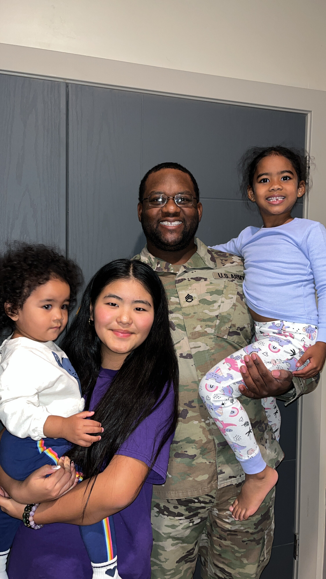 U.S. Army soldier in camouflage uniform poses with three young girls indoors, smiling together.