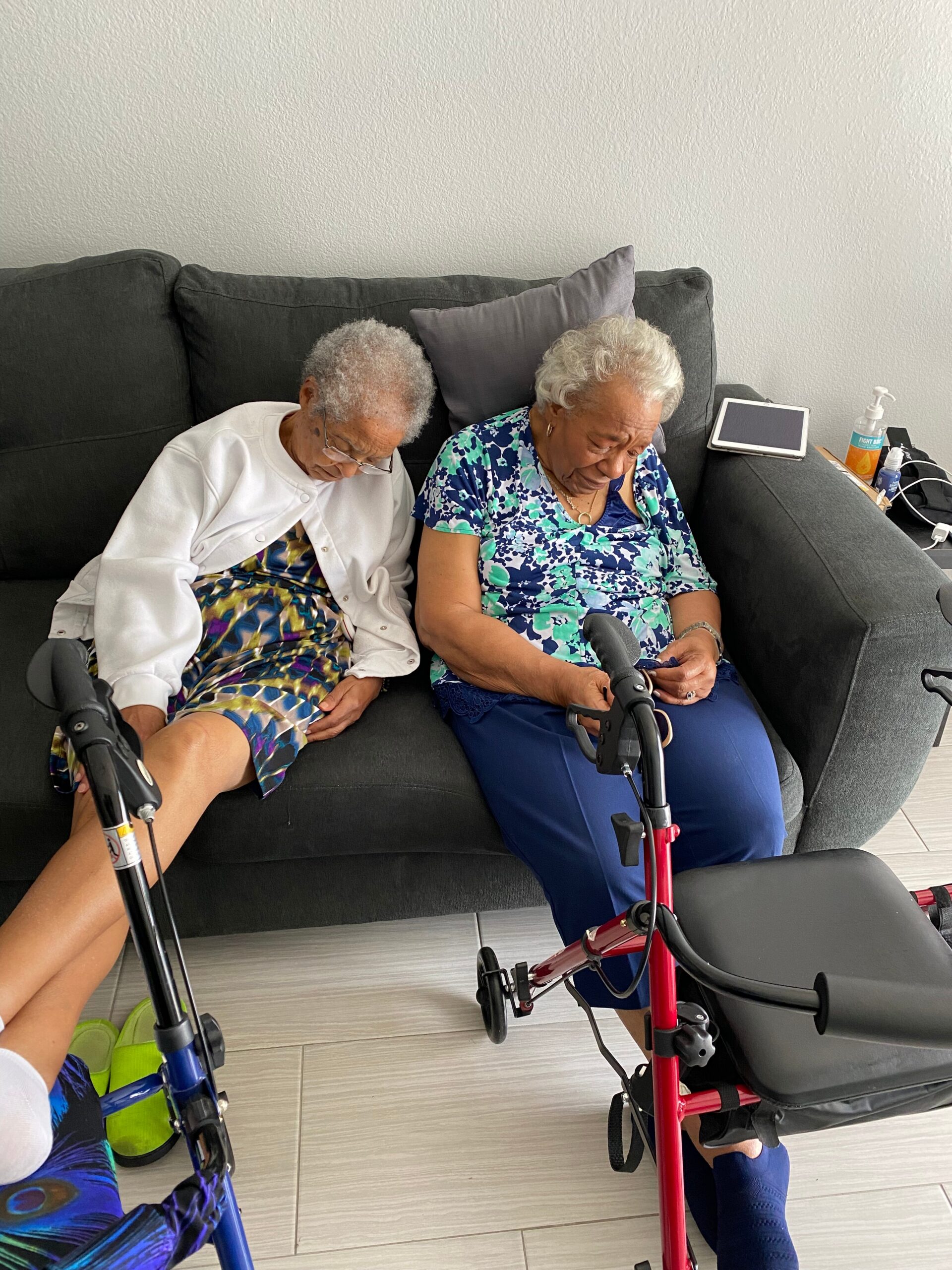 Two elderly women sit on a dark sofa at home, with mobility walkers in front and a tablet resting on the arm of the sofa nearby.