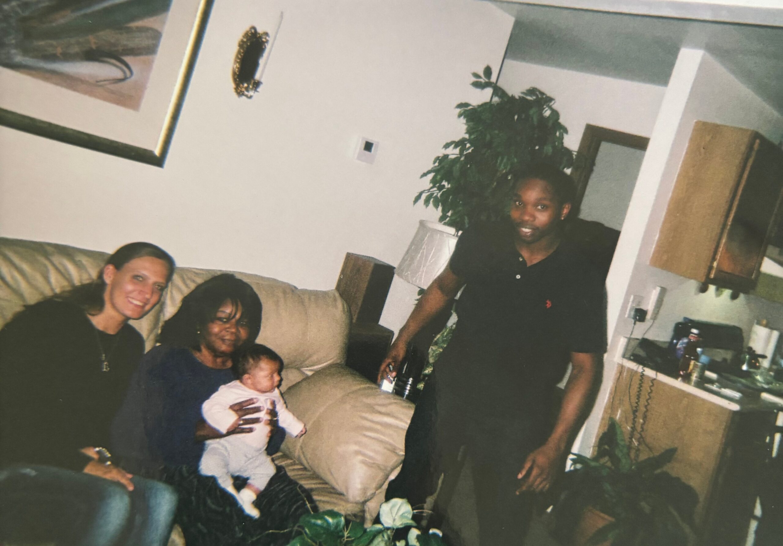 Family scene in a living room: adults on a couch with a baby, a standing woman nearby, houseplant and furniture in view.