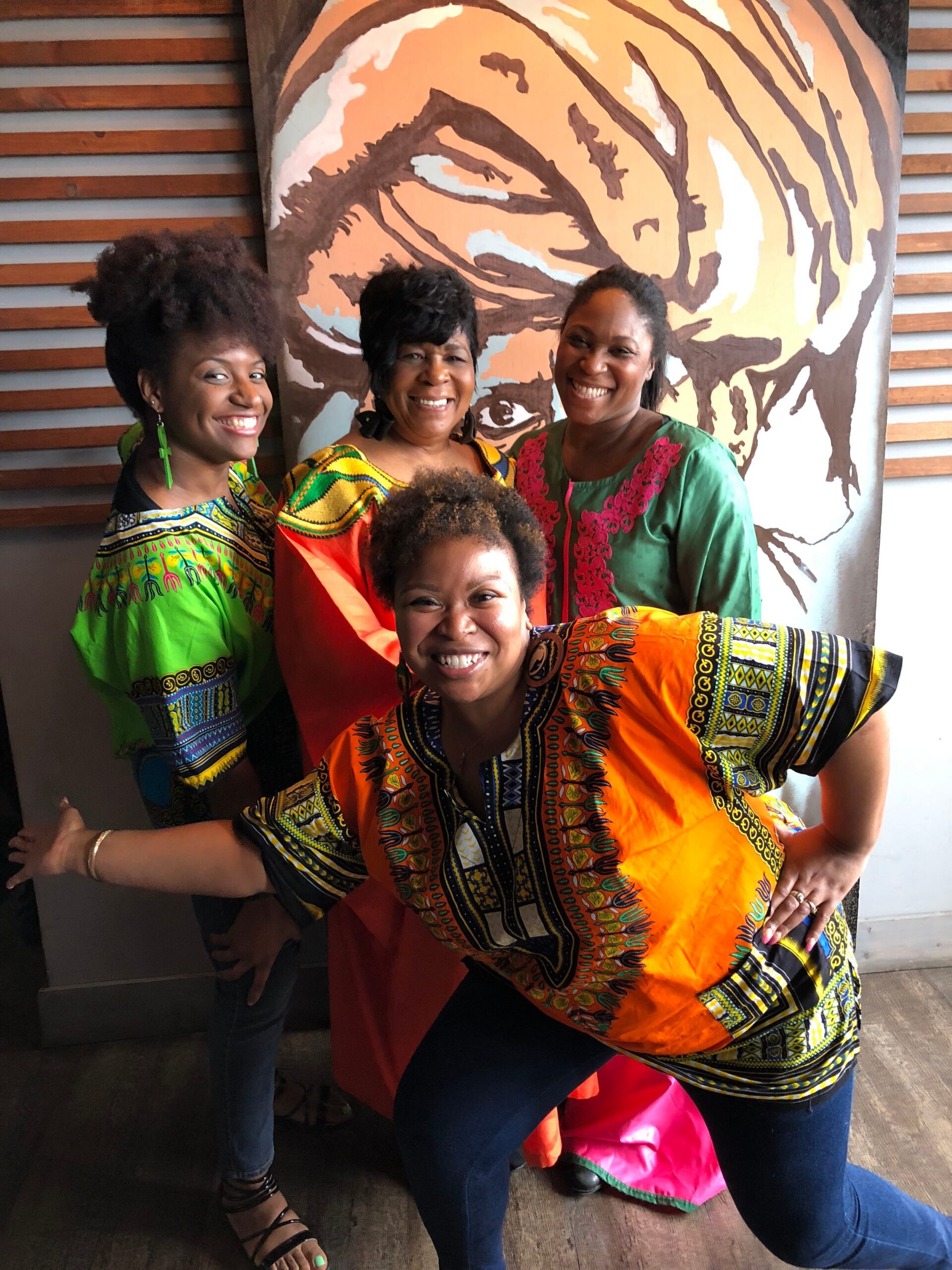 Four women wearing vibrant African dashiki outfits smile and pose together indoors in front of a large tiger mural on the wall behind them.