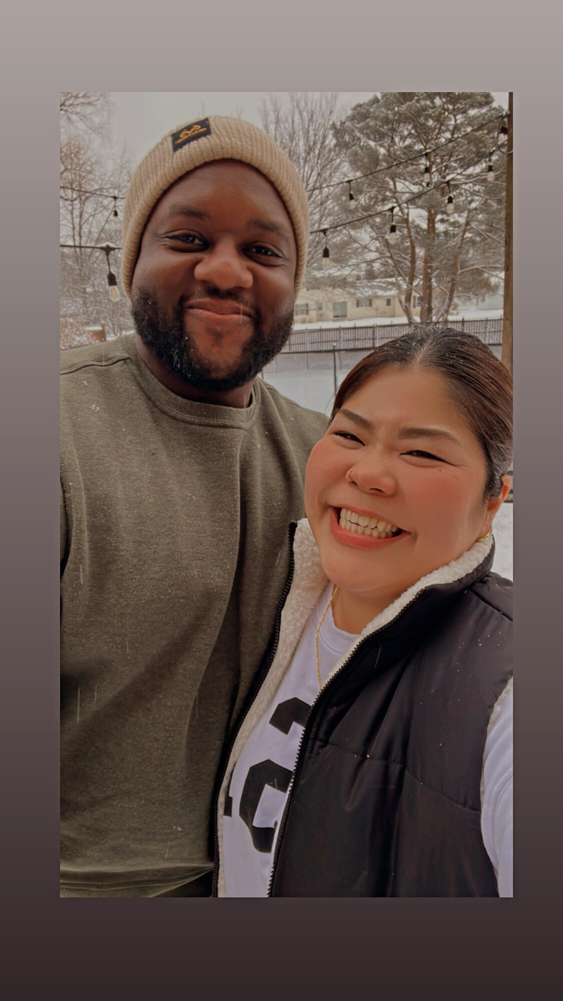 Two friends posing for a snowy selfie outdoors: a man in a beige knit beanie and olive sweater beside a woman in a black jacket, both smiling warmly with a winter backdrop of trees and string lights.