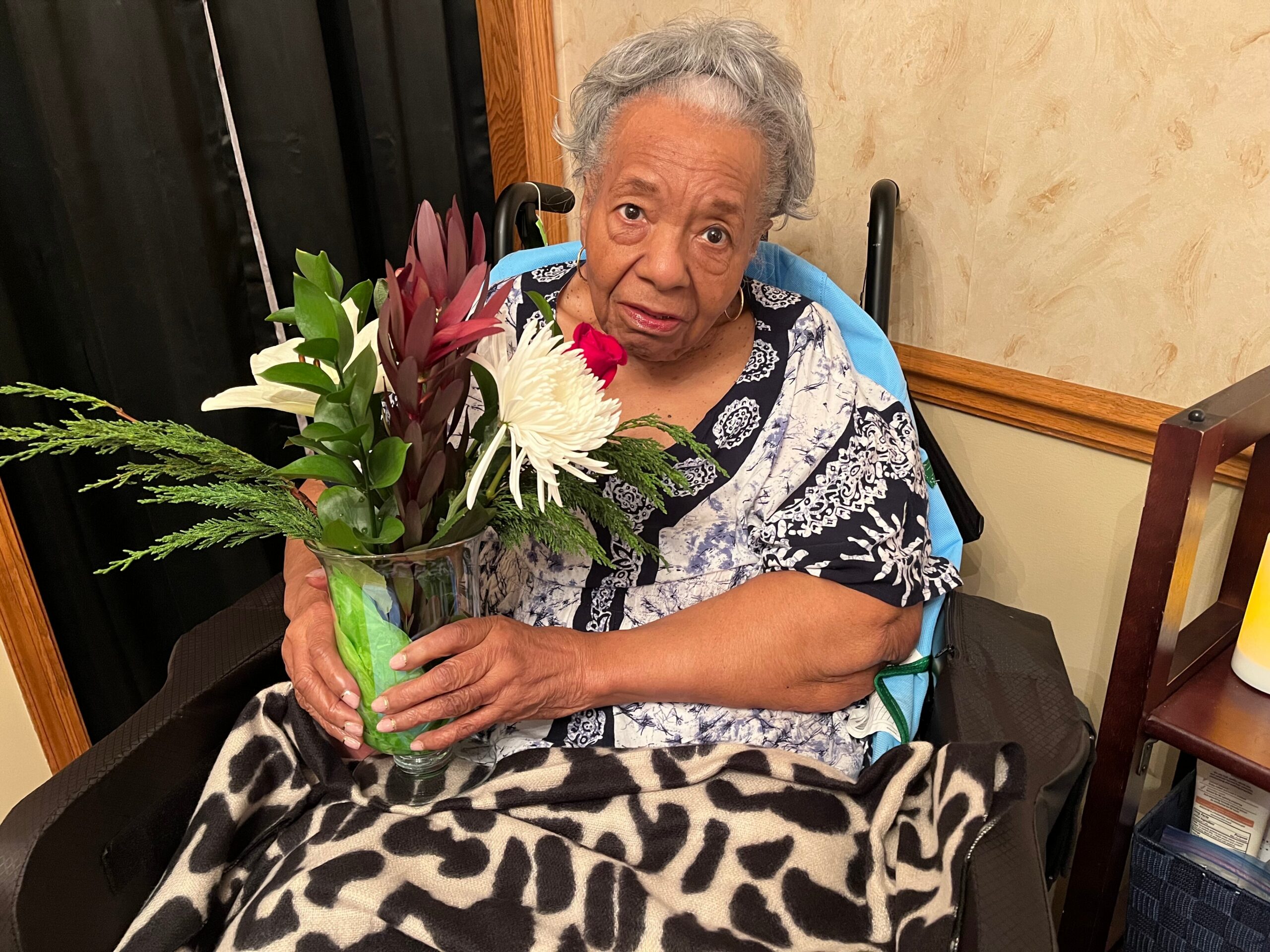 Elderly woman in a wheelchair holding a vase of mixed flowers, looking at the camera with a gentle smile.