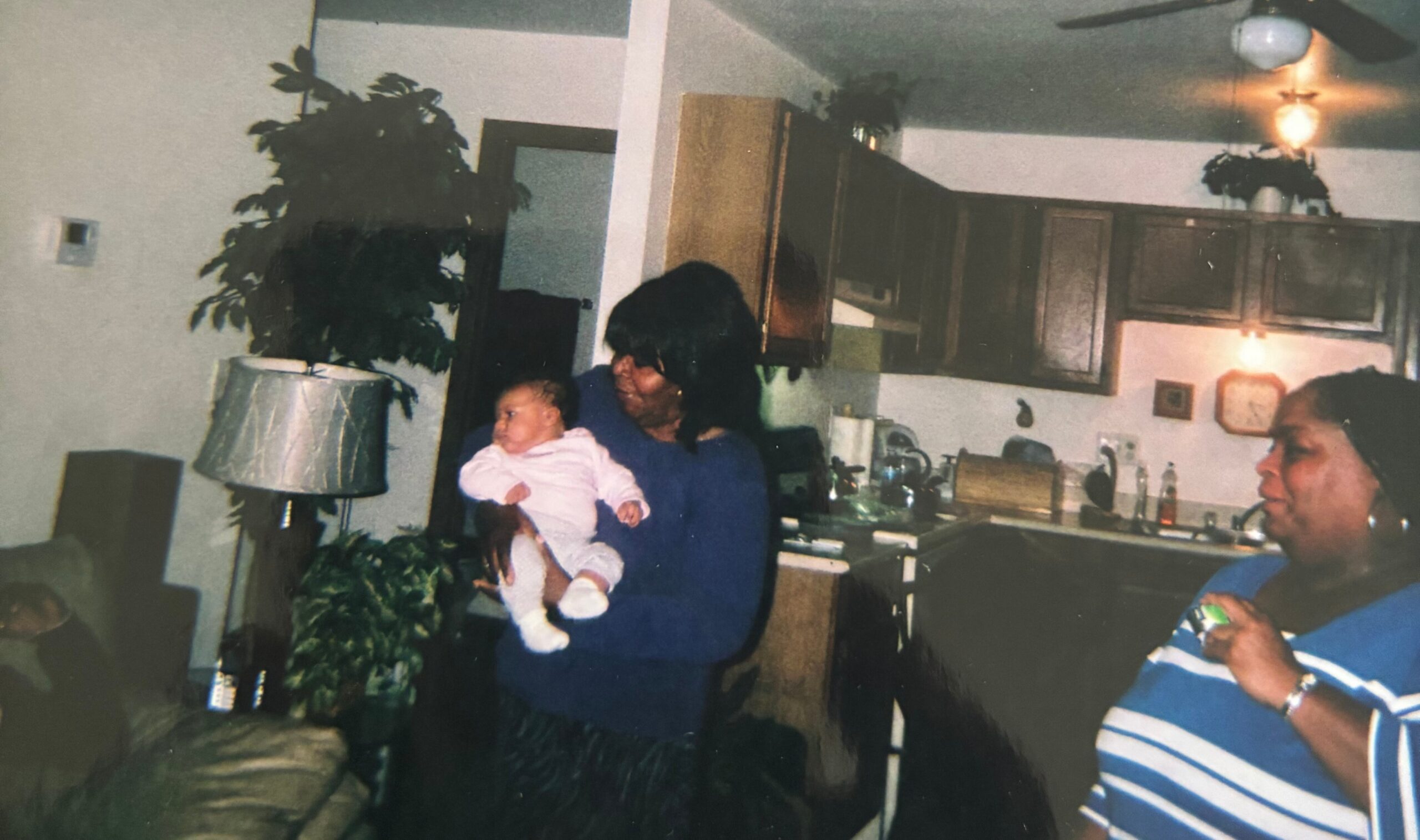 Woman in a blue outfit holding a baby in a dimly lit kitchen with dark cabinets and plants in the background.
