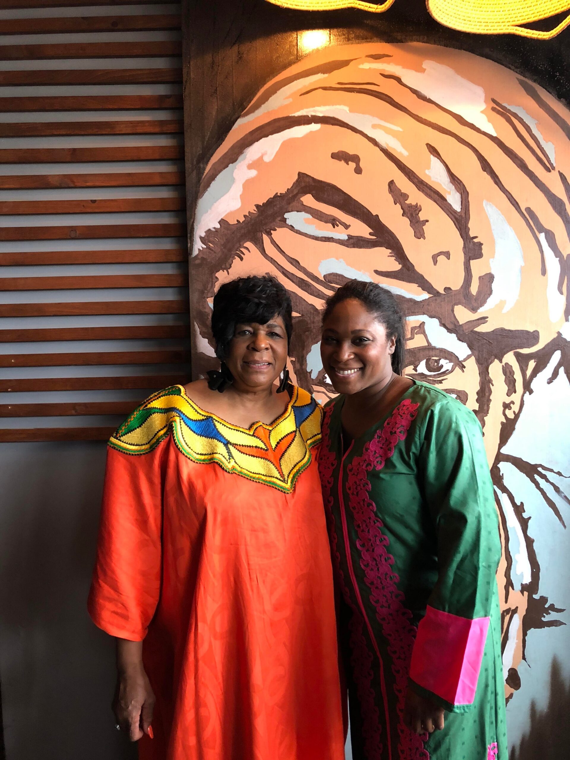 Two women in colorful African-print dresses pose together in front of a tiger mural with an eye-catching orange-and-brown design.