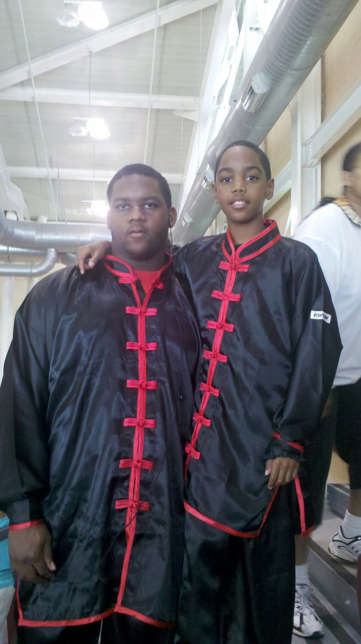 Two young people wearing matching black satin kung fu uniforms with red trim, standing side by side in an indoor facility.