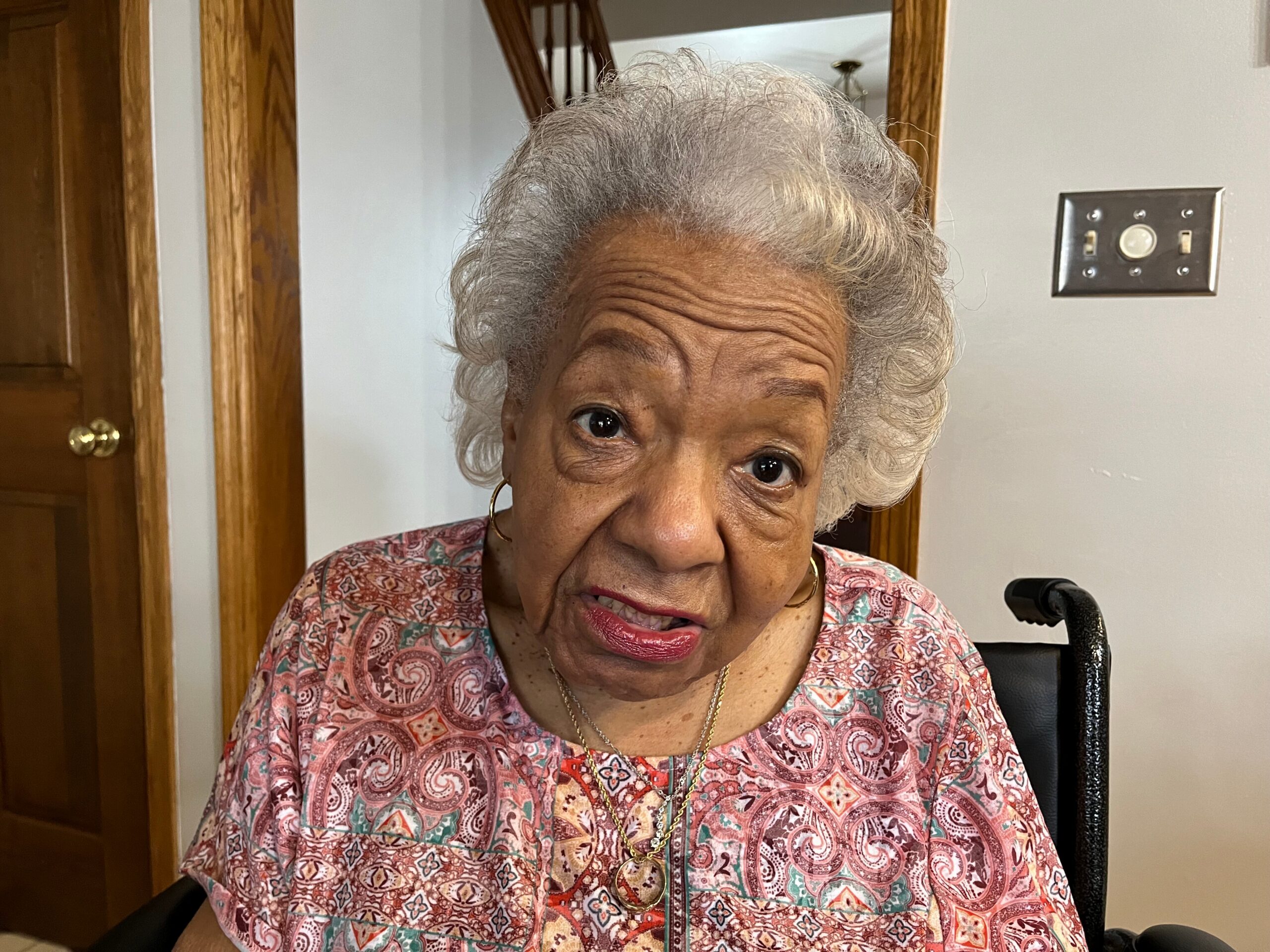 Older woman with gray hair and pink lipstick wearing a patterned blouse, seated indoors by a doorway.