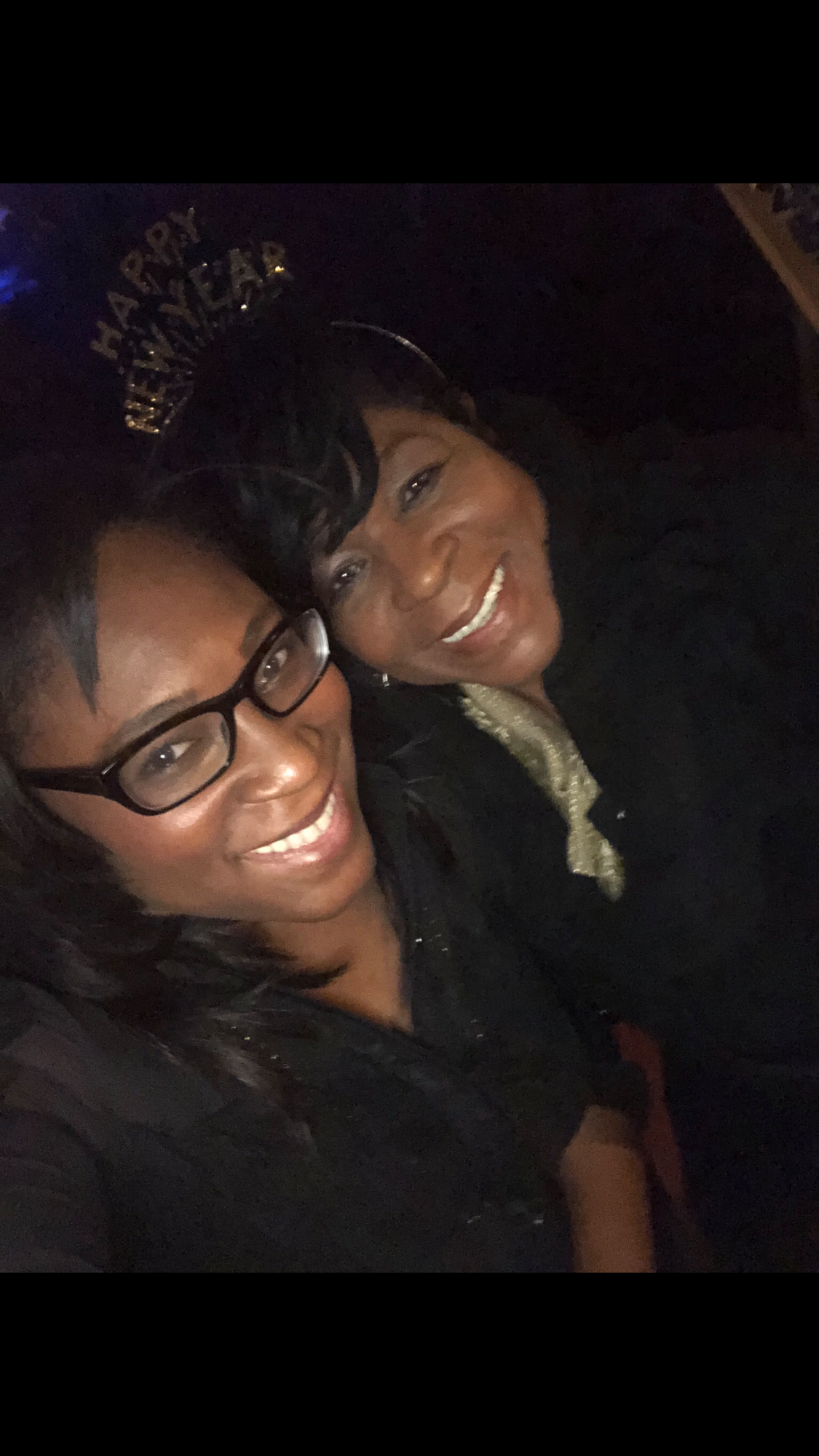 Two women smiling for a selfie in a dark setting; one wears glasses, the other wears a light scarf and a glittery headband that says 'Happy Birthday' or similar.