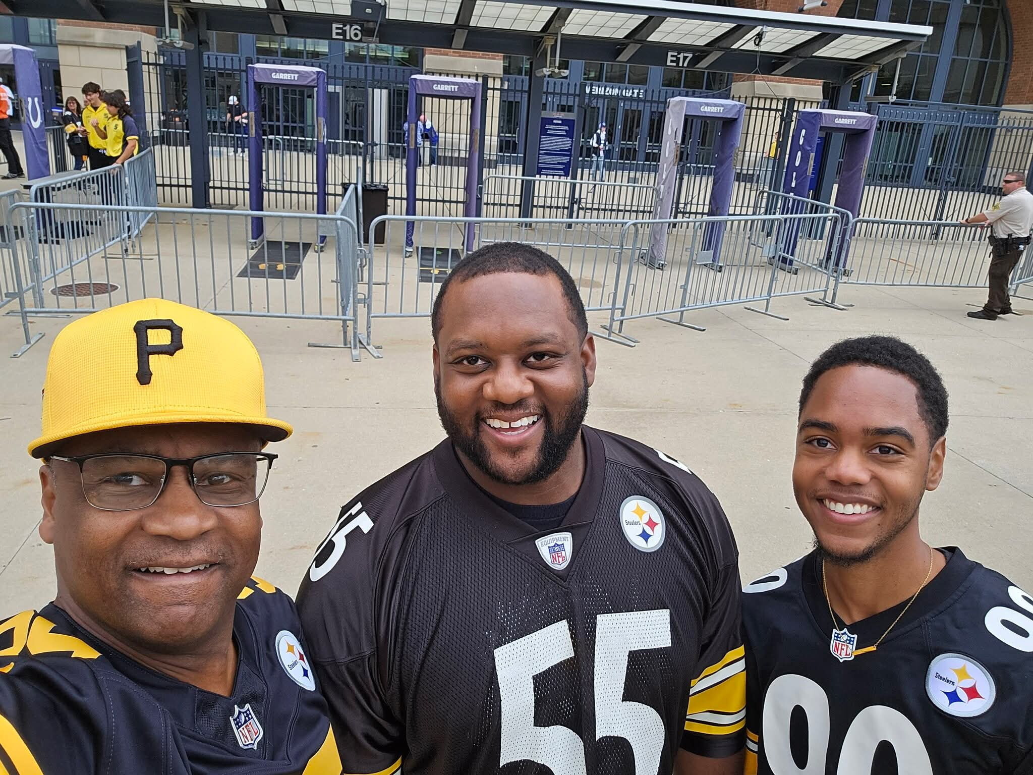 Three Steelers fans pose for a selfie outside a stadium, with metal barricades and purple security gates in the background.
