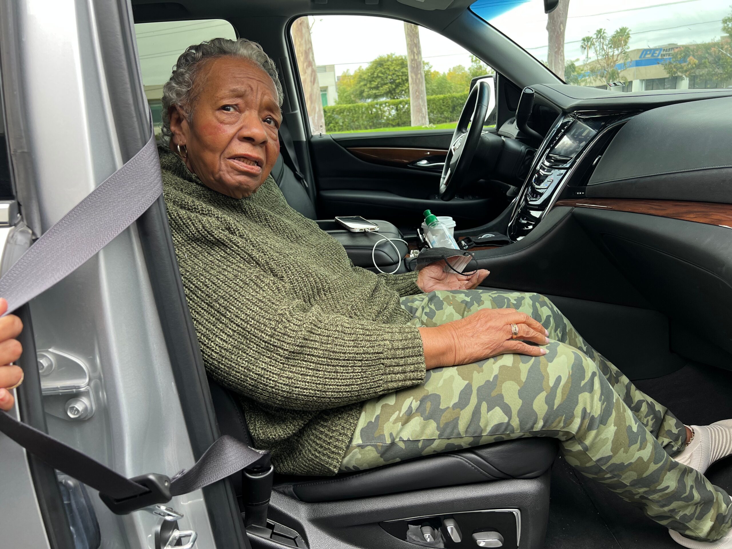 Old woman seated in a car passenger seat, wearing a green sweater and camouflage pants, looking toward the camera with a concerned expression while strapped in.