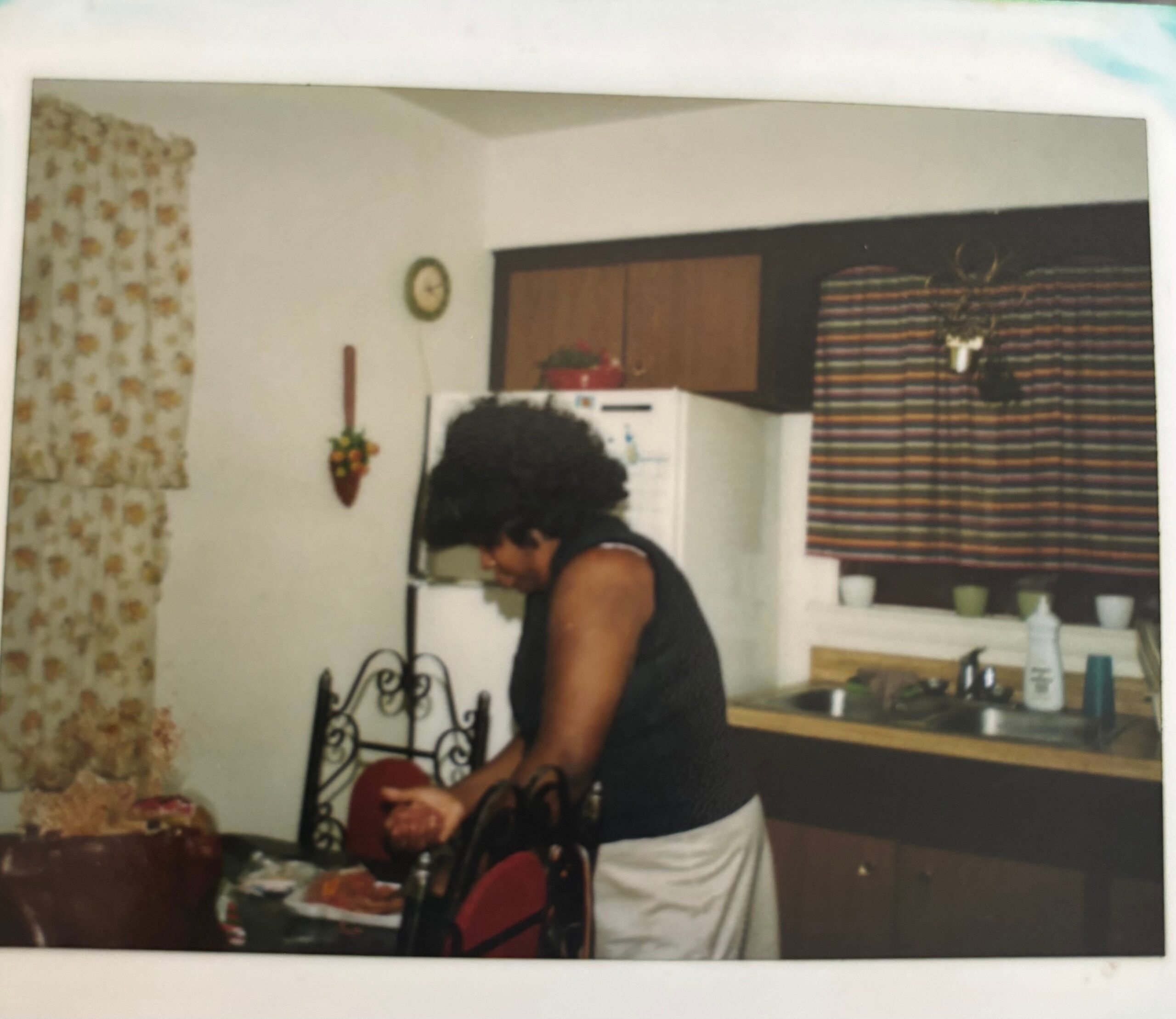 Woman wearing a sleeveless top stands at a kitchen counter with food items spread out, floral curtains to the left and a colorful striped curtain to the right, clock on the wall behind.