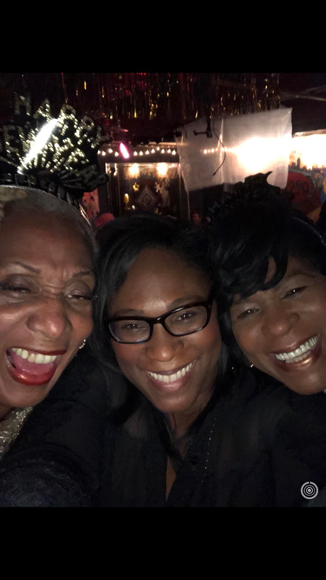 Three women taking a close-up selfie, smiling broadly at a festive nighttime event with string lights in the background