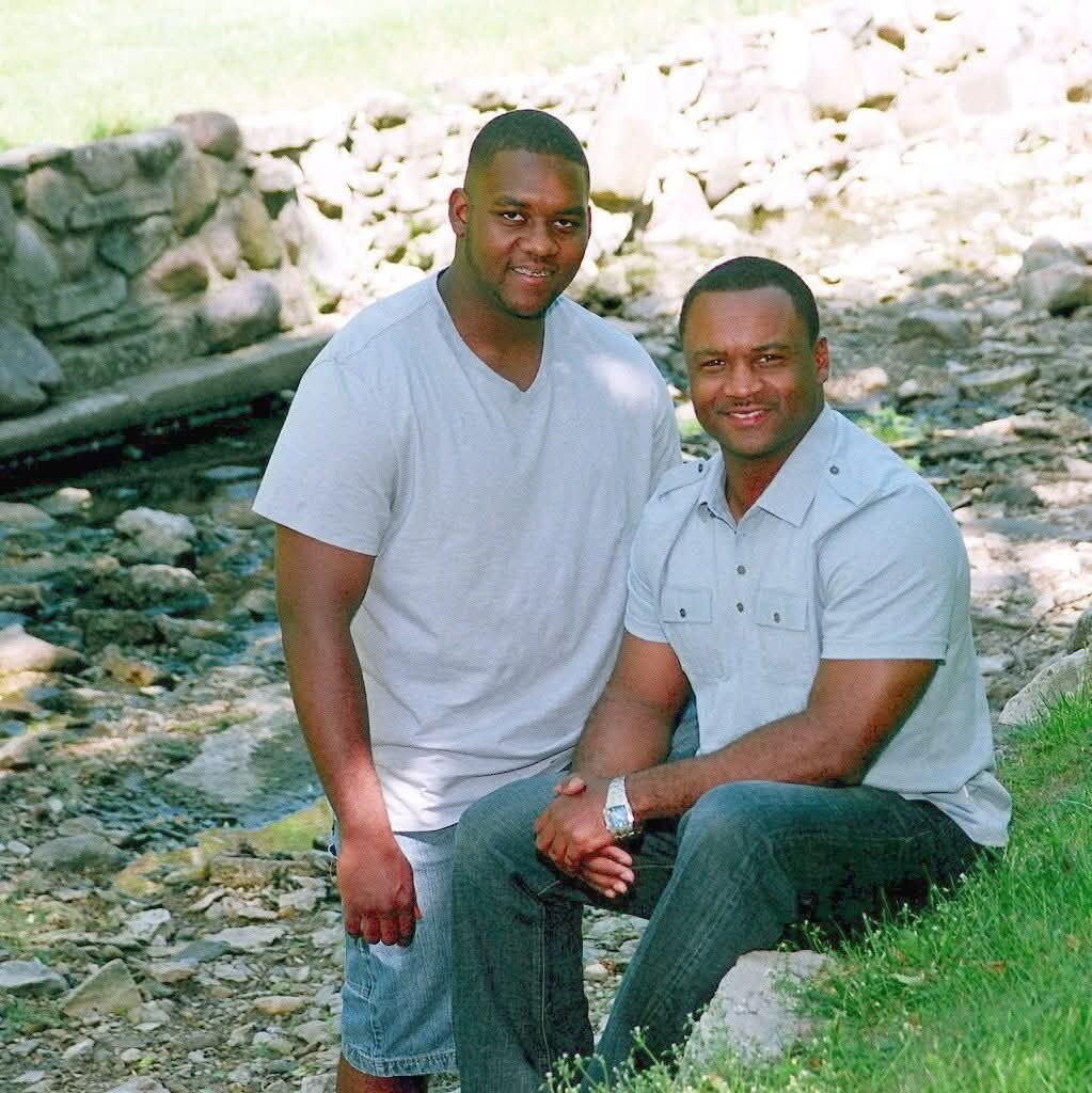 Two men pose outdoors by a rocky creek bed; one stands, the other sits on a rock and they smile at the camera.