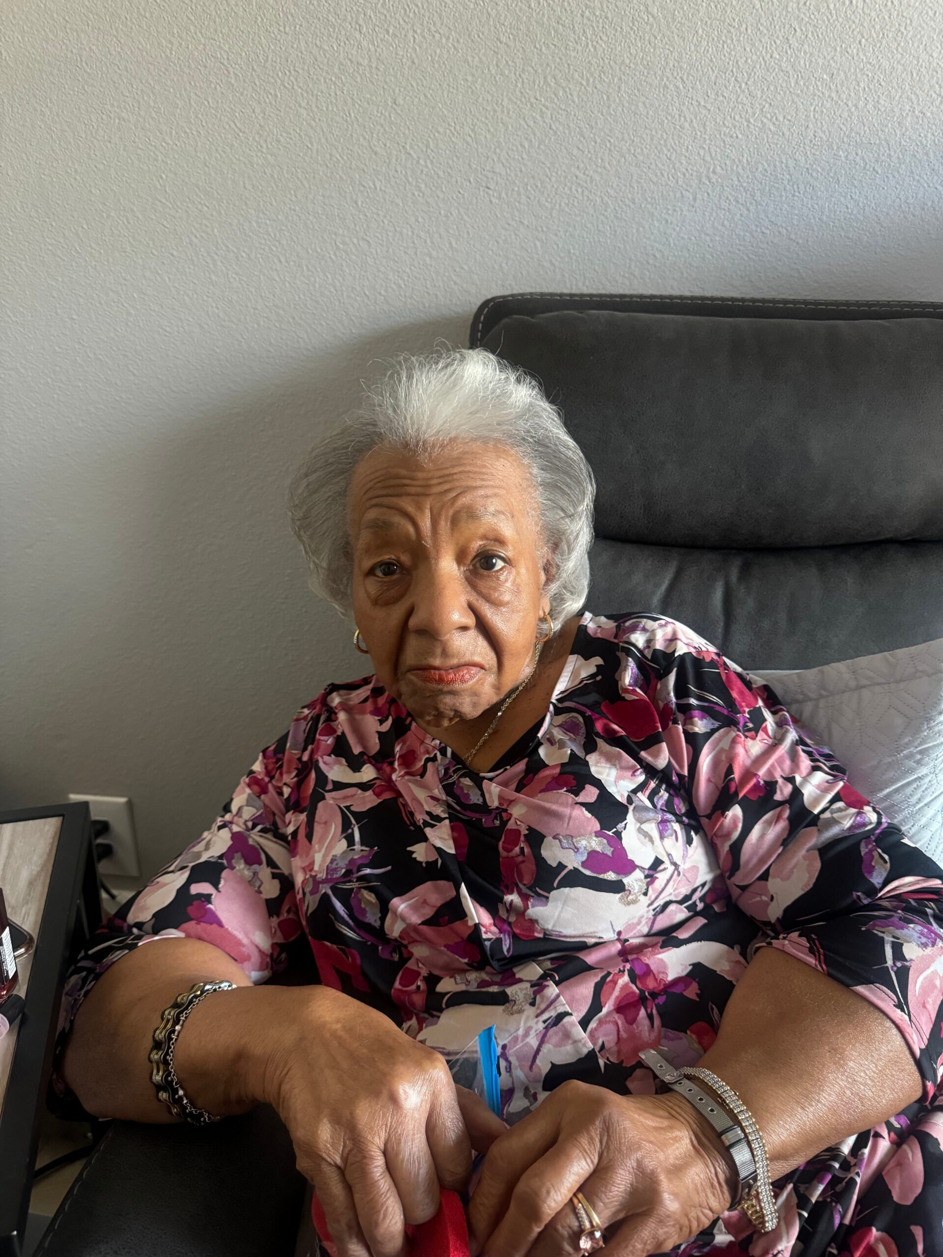 Elderly woman with short white hair sits on a dark sofa, wearing a pink and purple floral blouse, hands resting on a table.
