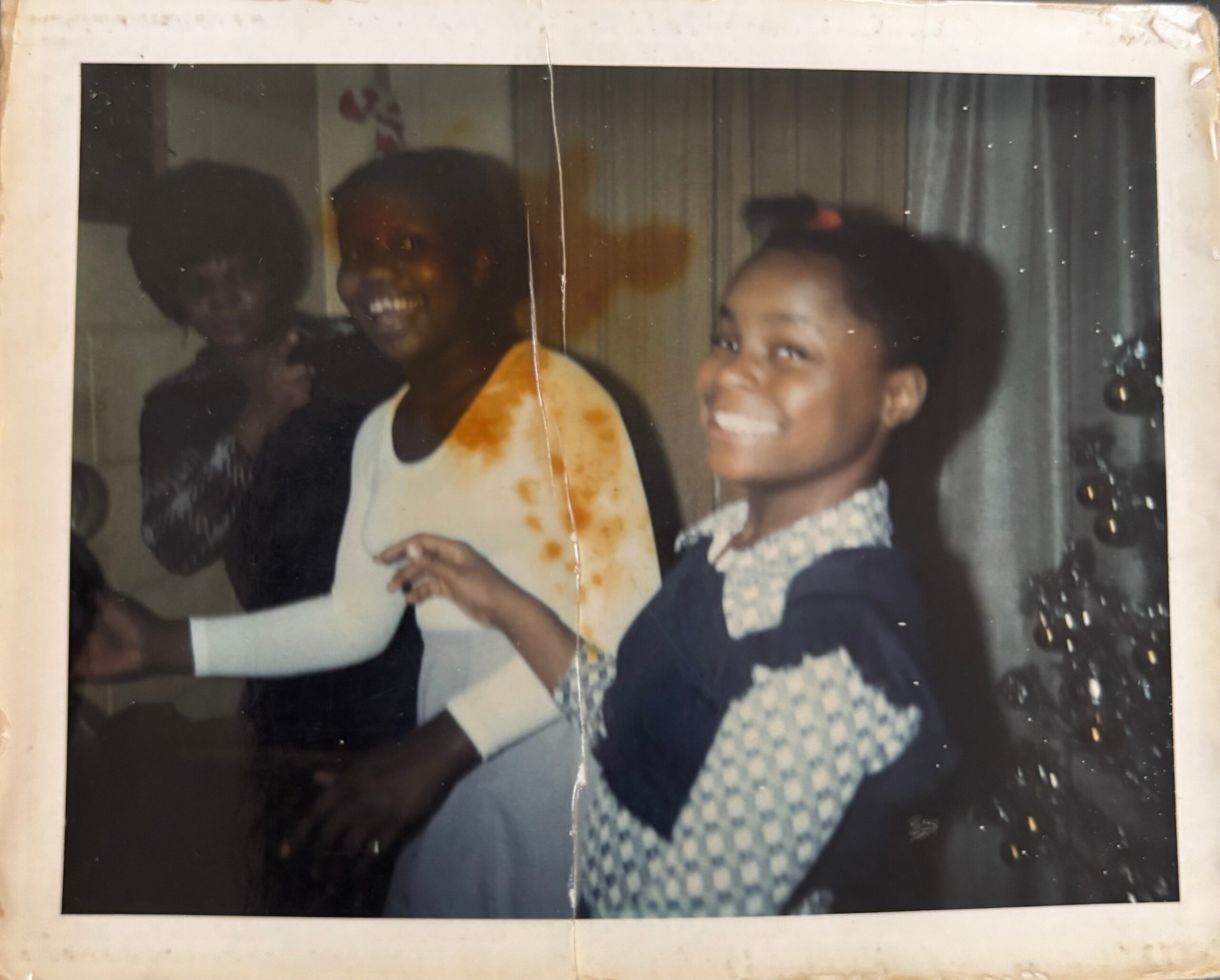 Two smiling young girls pose indoors for a photo; one wears a light stained shirt, the other a dark dress with lace collar.