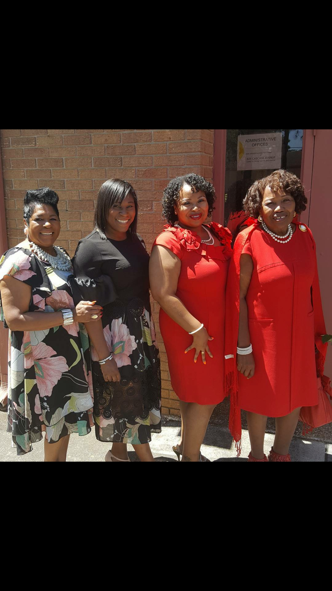 Four women stand side by side outside a brick building, posing for the camera in colorful dresses (two in red, one in black floral, one in black).