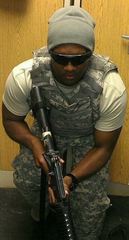 Soldier in camouflage vest and gray beanie crouching while aiming a rifle, wearing sunglasses.