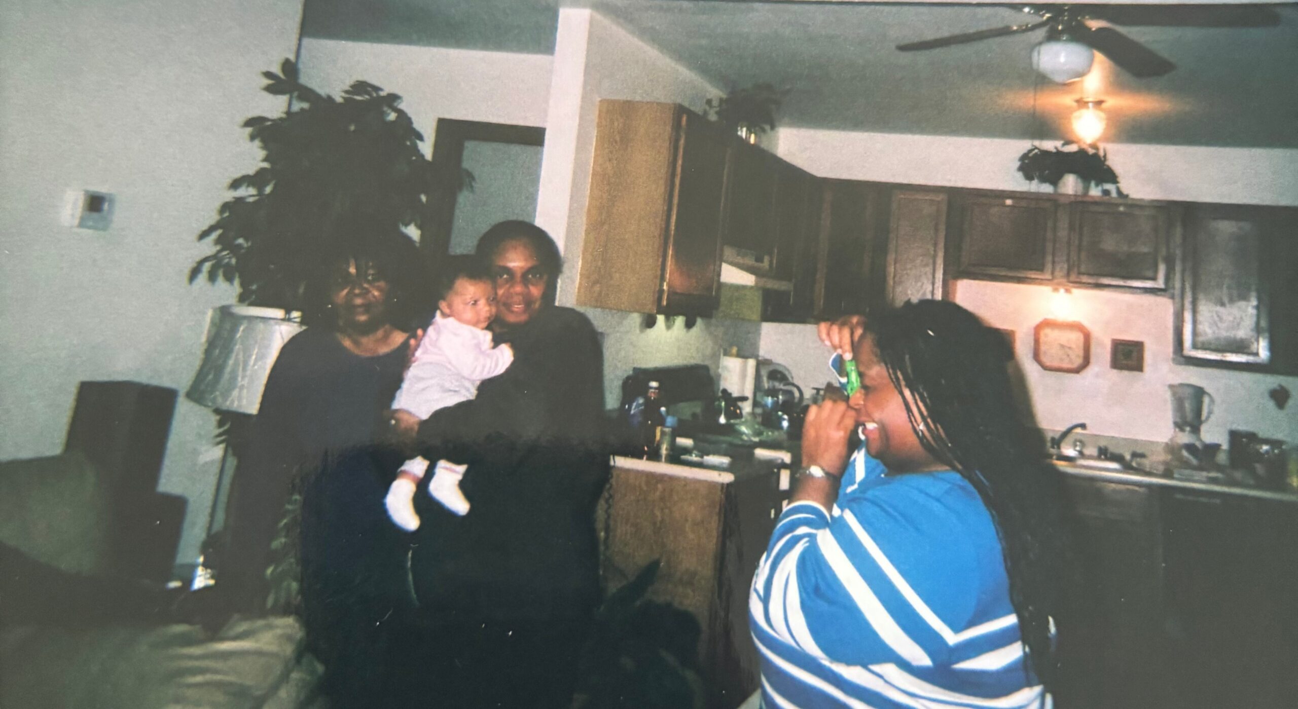 Family in a home kitchen: a woman holds a baby while another person stands nearby, and a third person in a striped blue shirt faces away on the right.