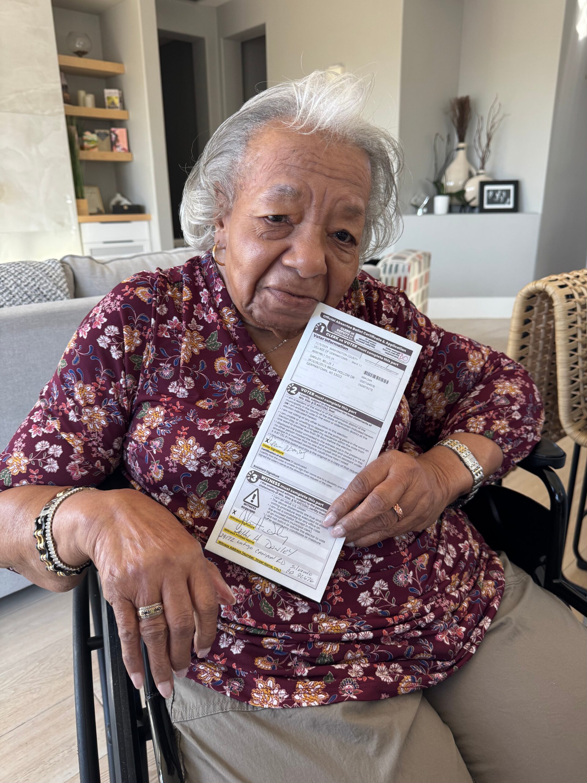 Elderly woman in a floral maroon blouse sits in a wheelchair, holding a voter form toward the camera in a bright living room.