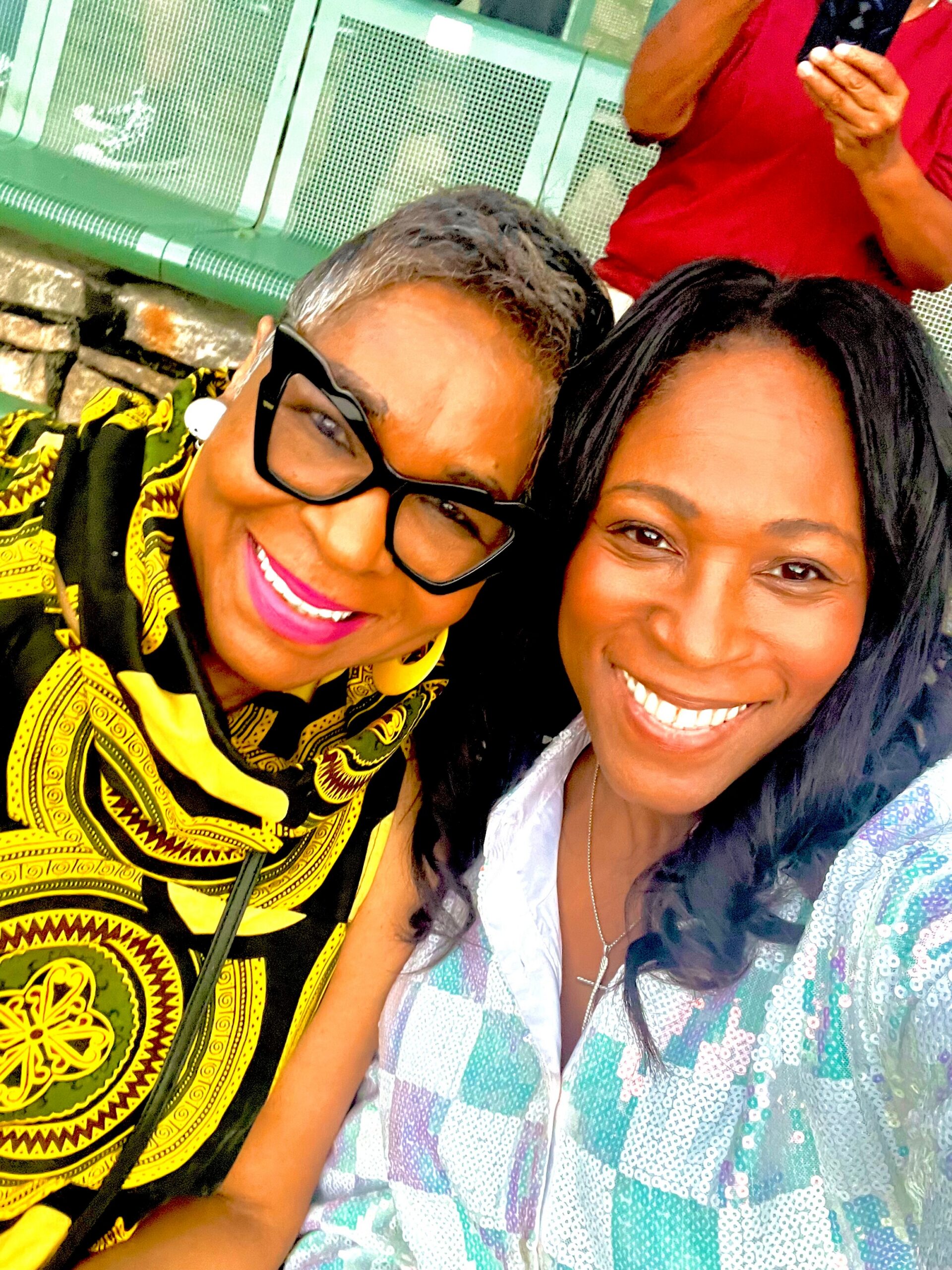 Two women taking a close-up selfie, smiling at the camera outdoors; one wearing a bright yellow and black patterned outfit with large glasses, the other in a light blue patterned blouse.