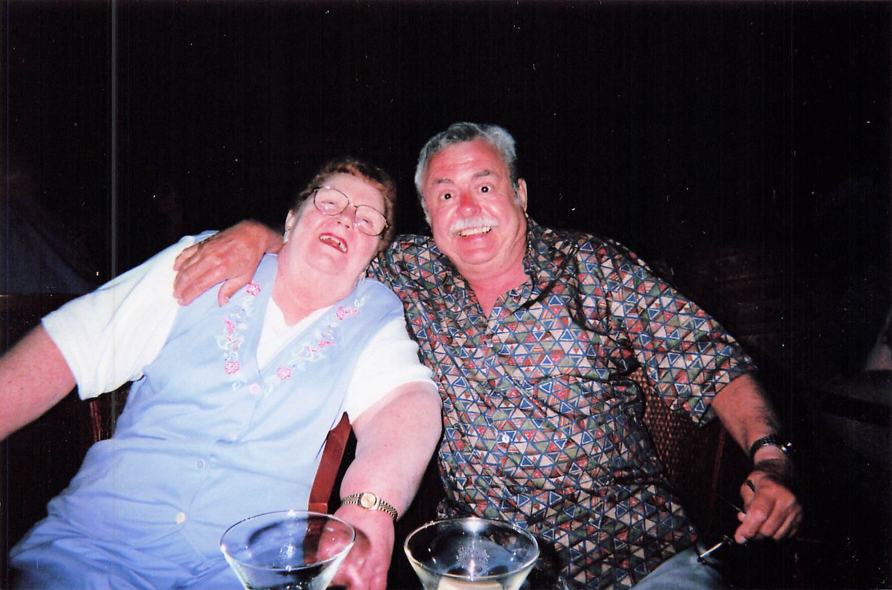 An elderly couple sits close together, smiling at the camera, arms around each other, with two cocktails in the foreground during a nighttime gathering.