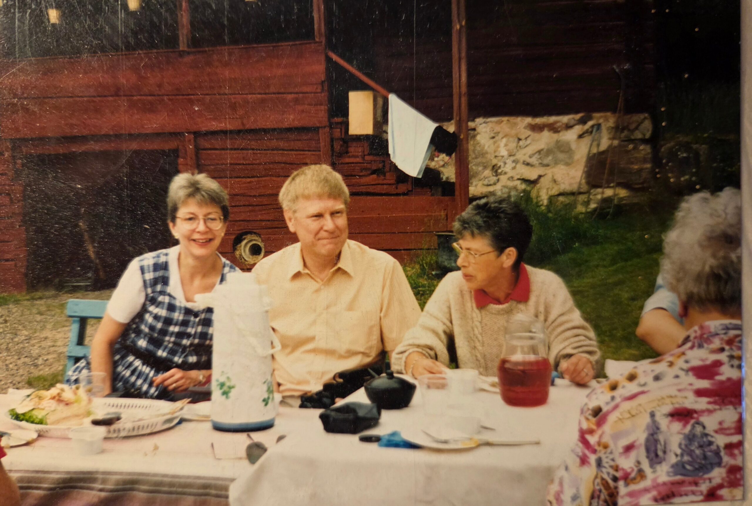 Group of people seated at a outdoor picnic table, smiling and talking during a backyard meal with drinks and plates on the table.