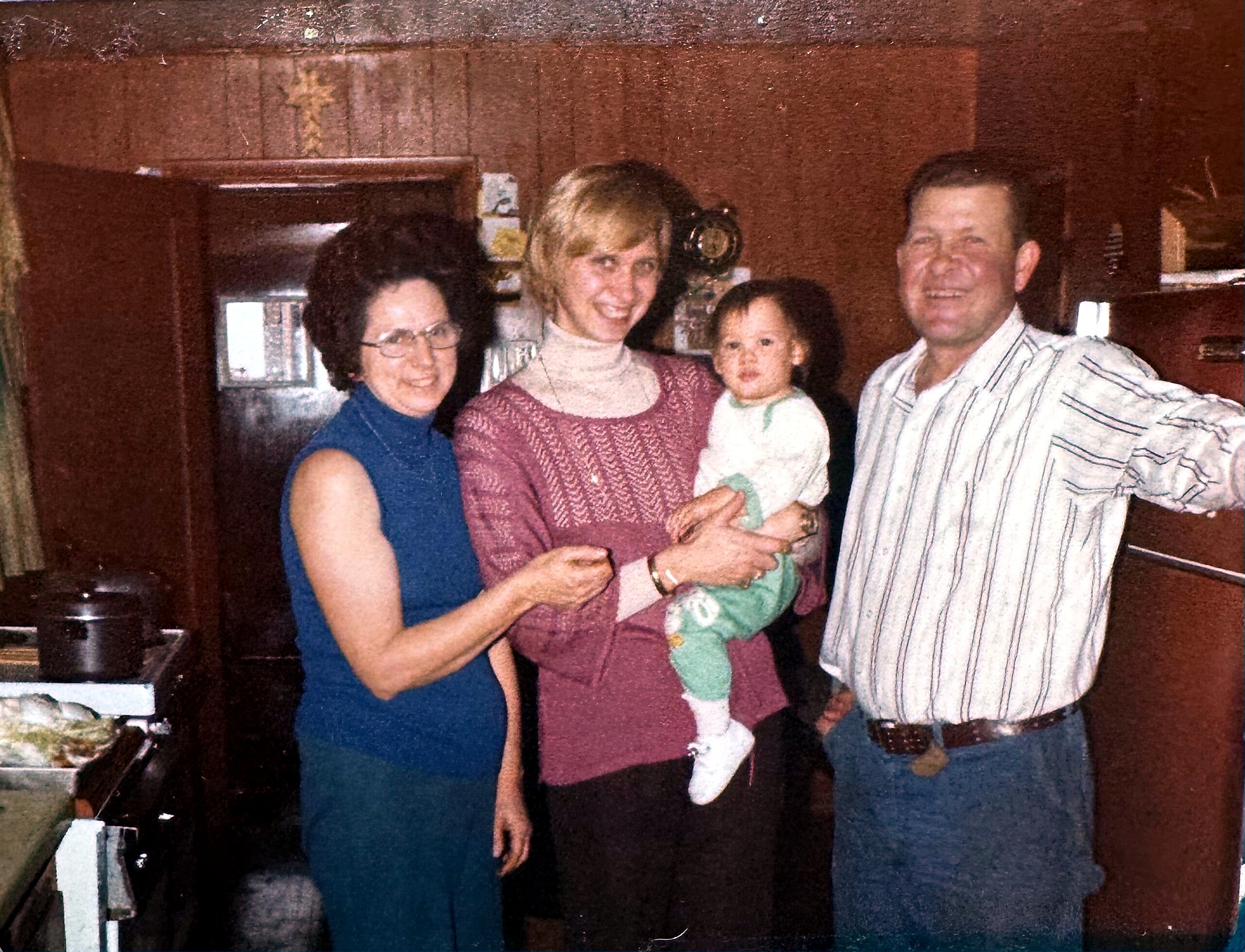 Four people—two women, a man, and a child—smile together in a kitchen with dark wood cabinets in the background.
