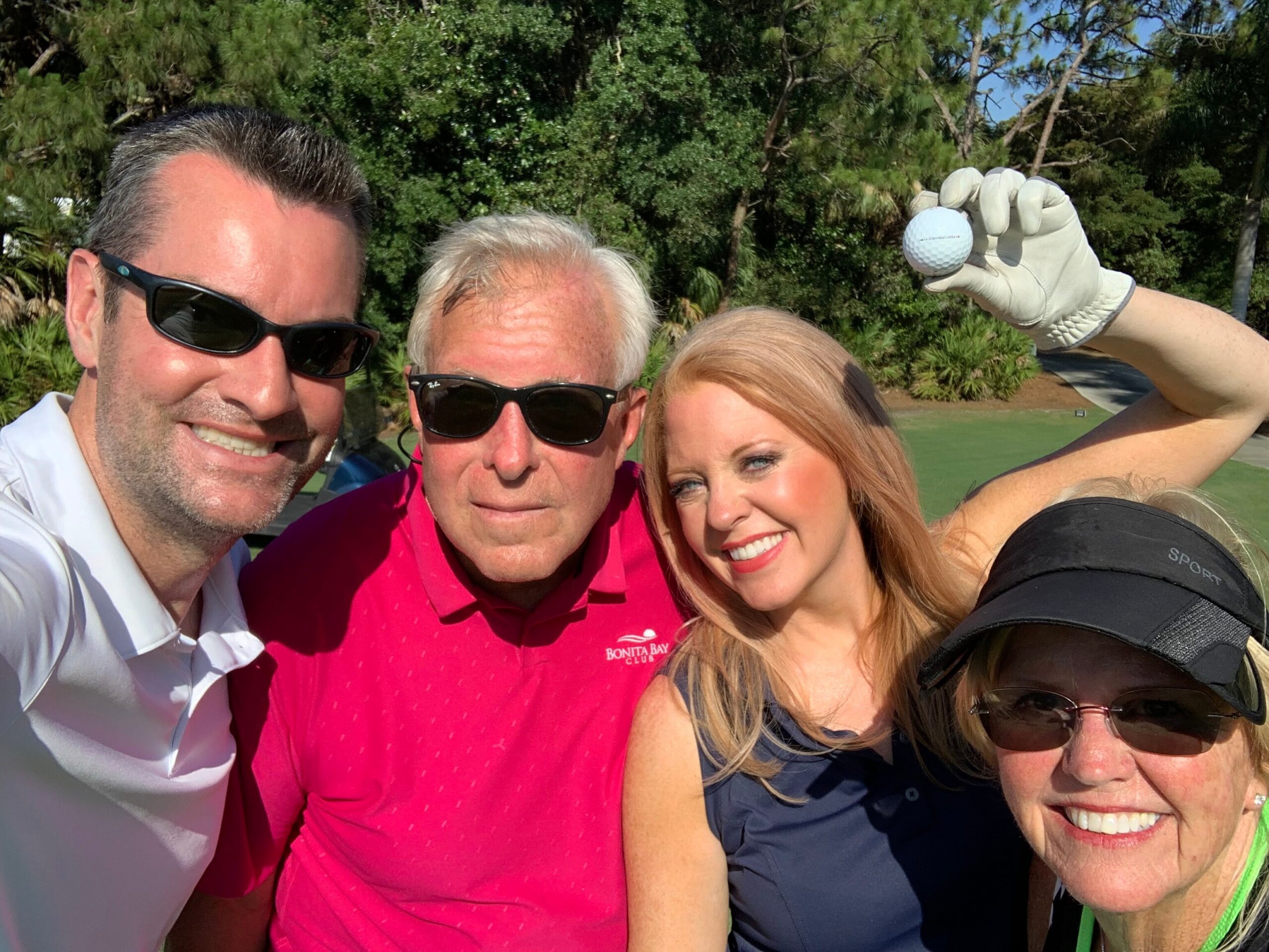 Four friends posing for a selfie on a golf course, one wearing a golf glove and holding a golf ball in the air.