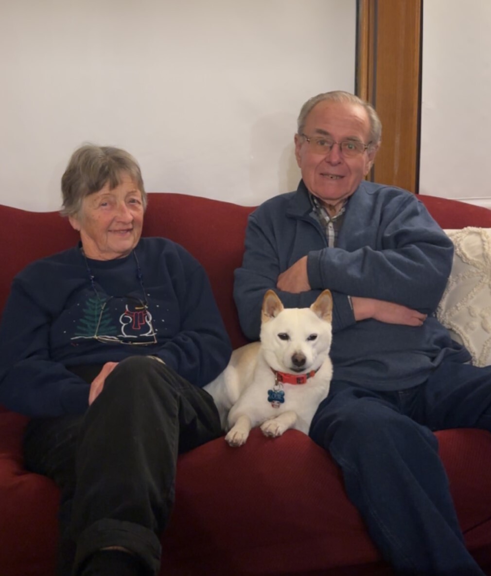 Elderly couple seated on a red sofa with a small white dog wearing a red collar between them.