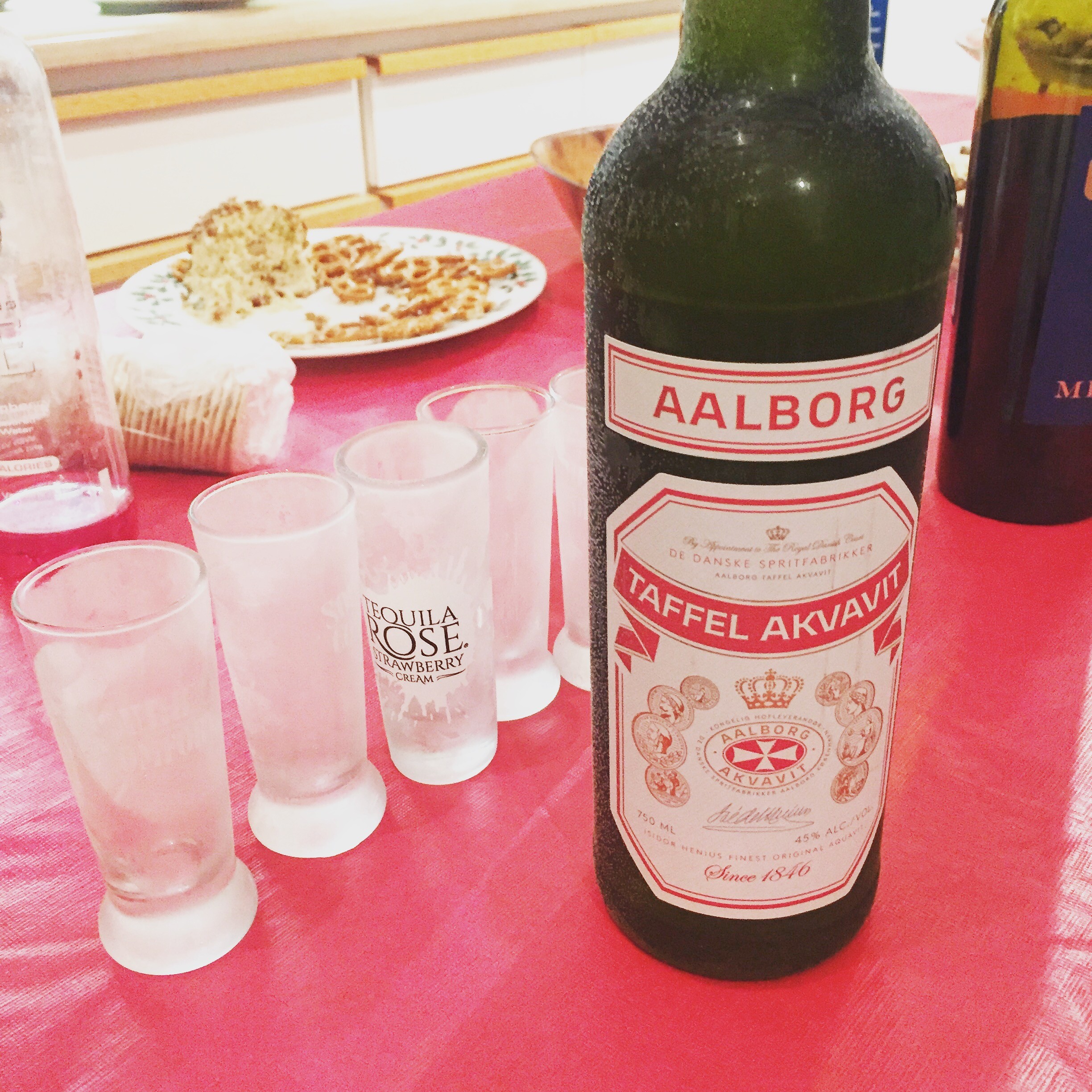 Bottle of Aalborg Akvavit on a red tablecloth, surrounded by shot glasses and a plate of snacks in the background