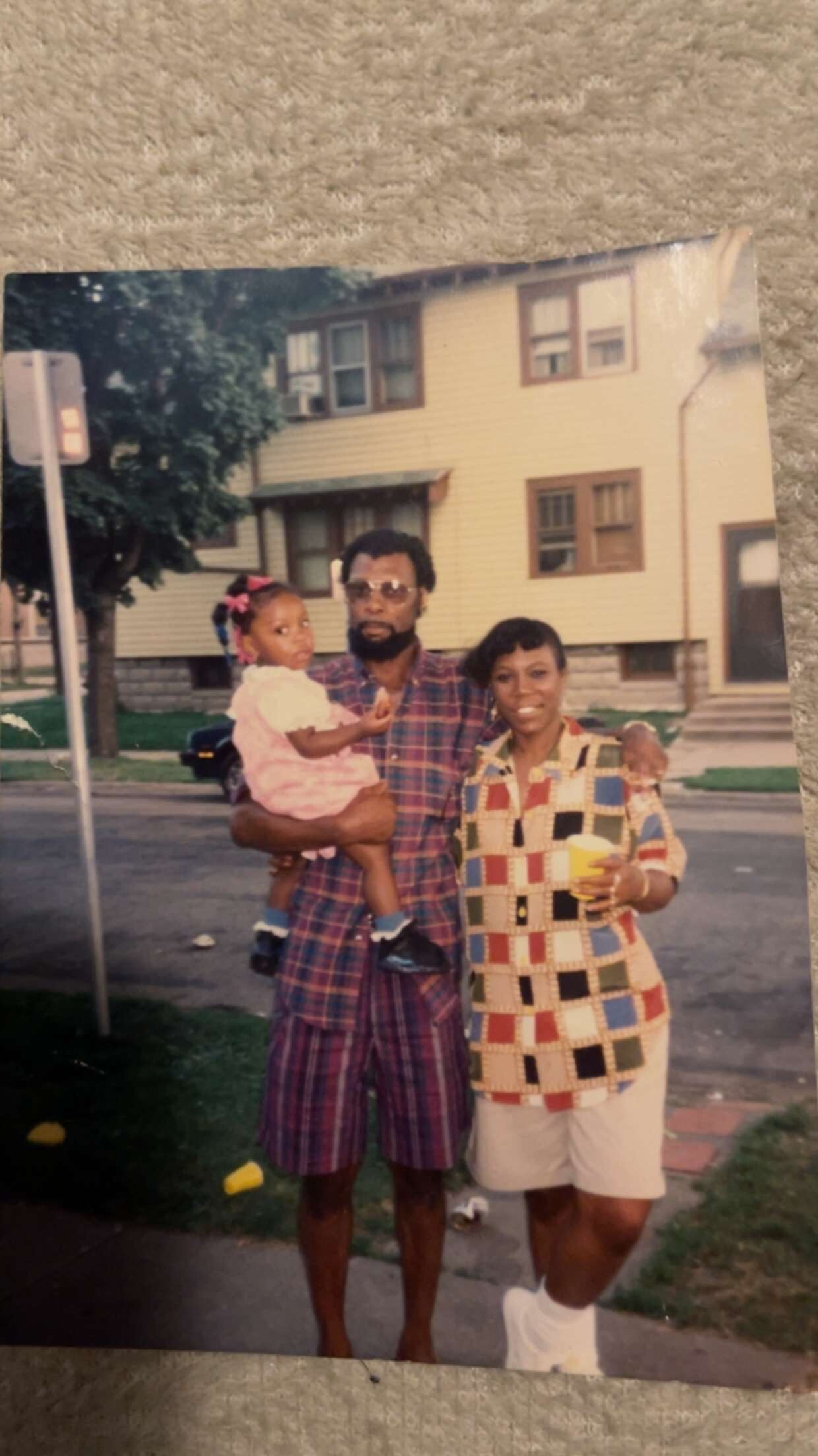 A man in a plaid outfit holds a young girl while a woman in a colorful patterned top stands next to them on a suburban street.