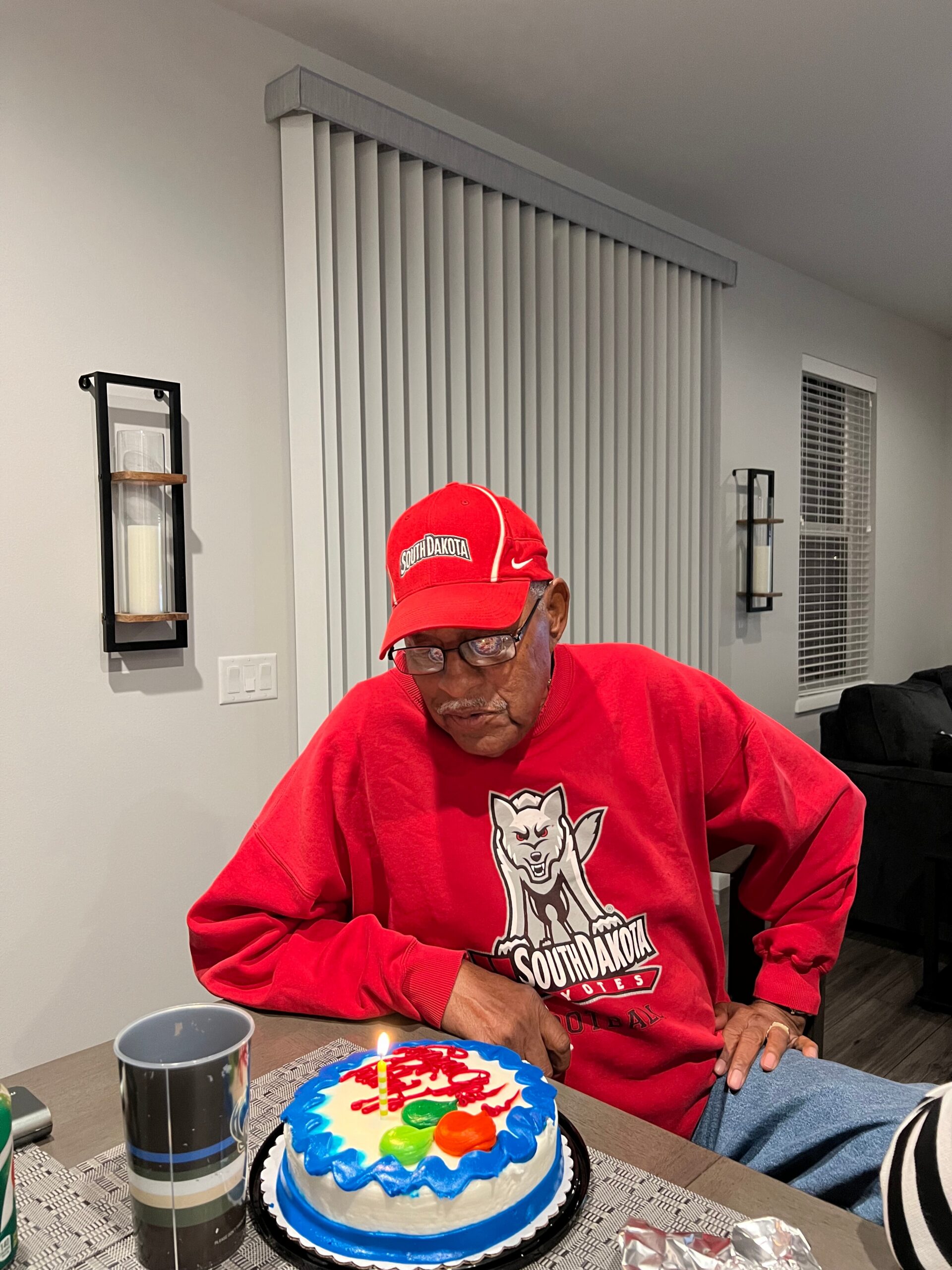 Older man in a red South Dakota shirt and cap sits at a table with a colorful birthday cake and a lit candle in front of him.
