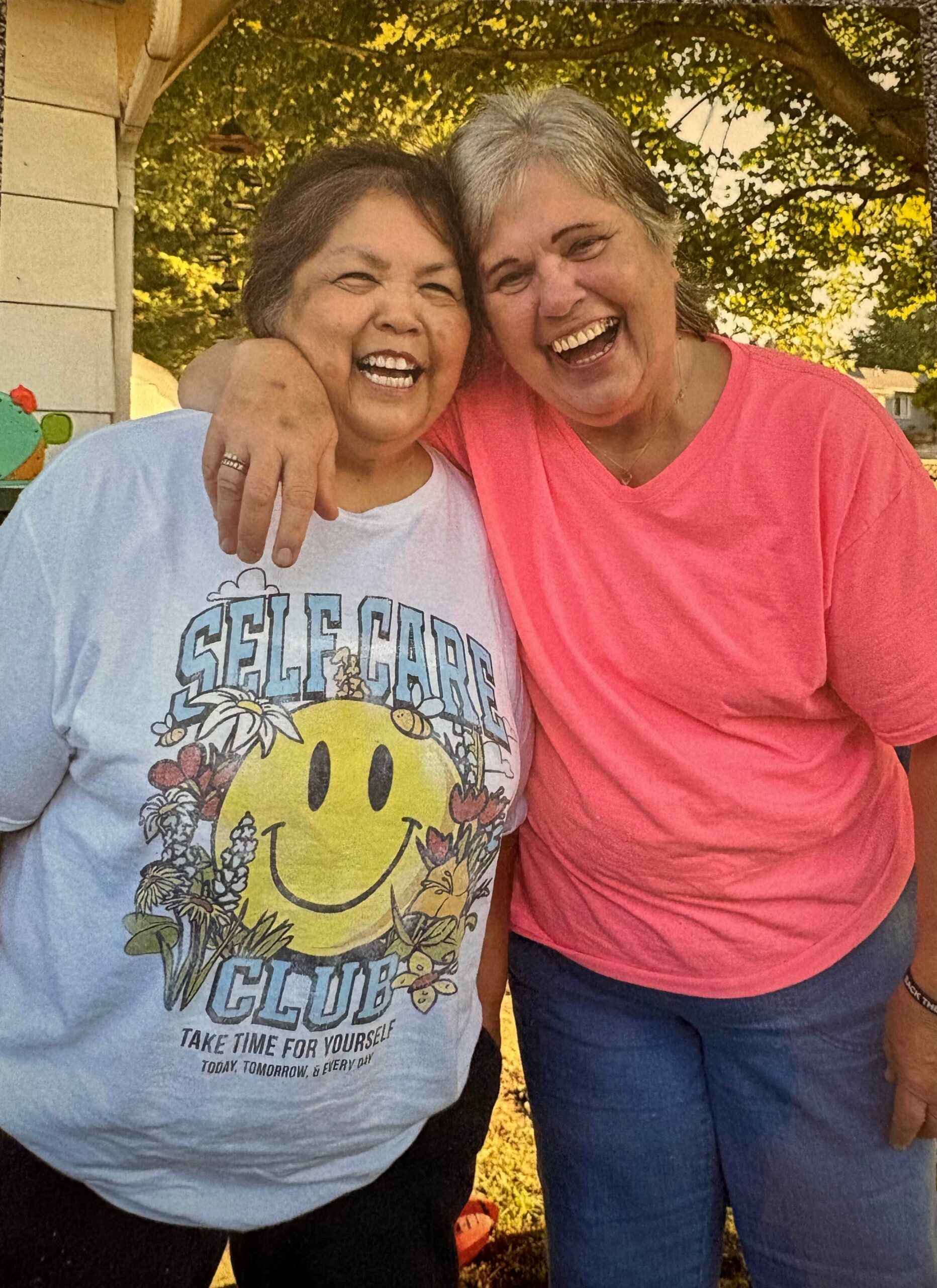 Two women outdoors, smiling with their arms around each other; one in a light blue graphic tee and the other in a pink shirt under leafy trees.
