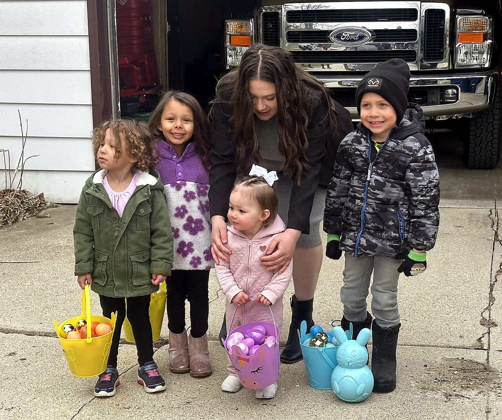 Children in coats hold colorful Easter baskets outside, smiling for a photo with a pickup truck in the background.