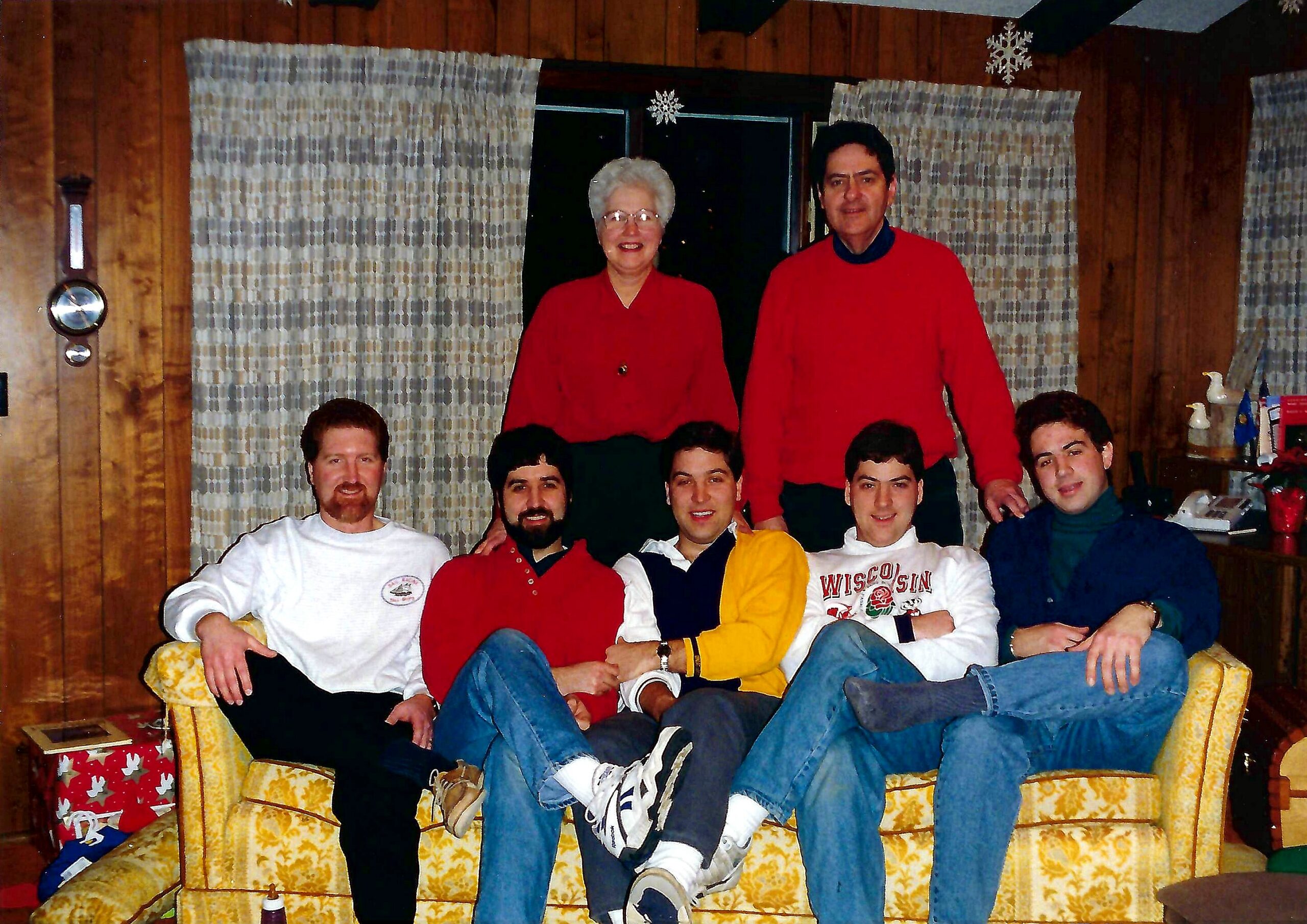 Group of people posing for a festive photo in a wood-paneled living room, some seated on a yellow patterned sofa and others standing behind, Christmas decorations visible in the background.