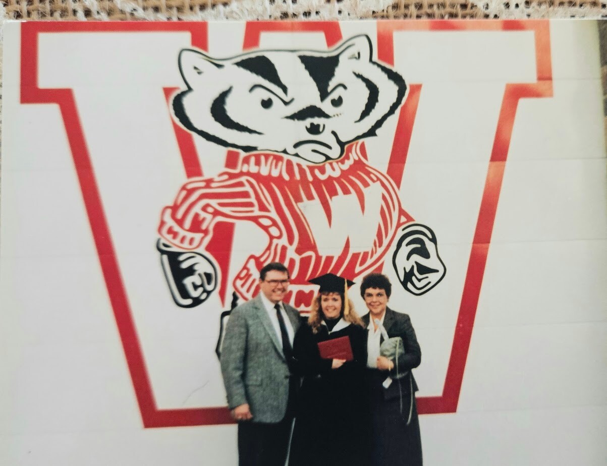 Three graduates—two women and a man—pose with diplomas in front of a large red W mascot mural (bulldog figure).