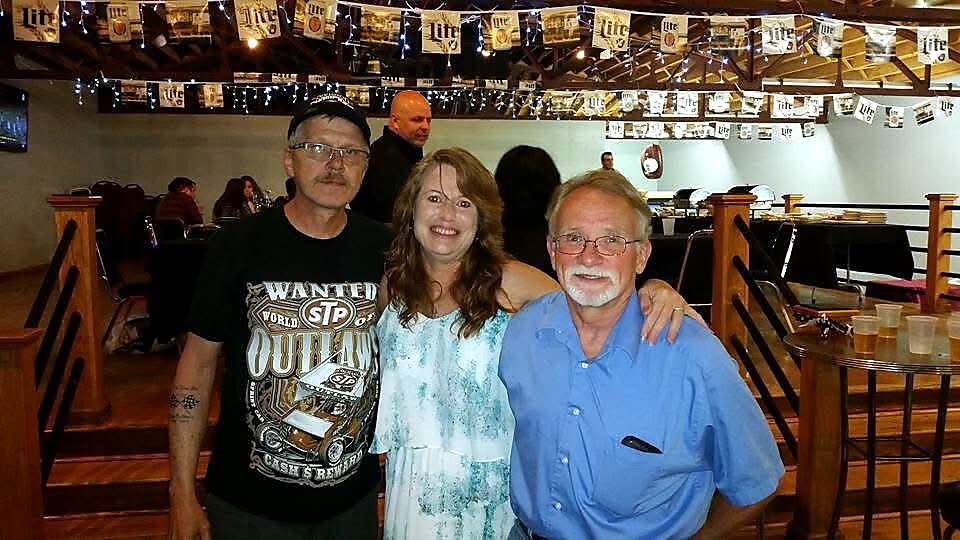 Three people posing for a photo in a bar: a man in a graphic black T‑shirt on the left, a smiling woman in a blue dress in the middle, and an older man in a blue shirt on the right.