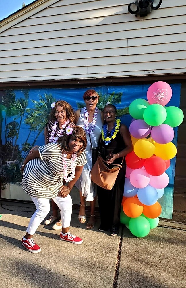 Four women wearing leis pose by a colorful balloon column at a tropical-themed outdoor party.