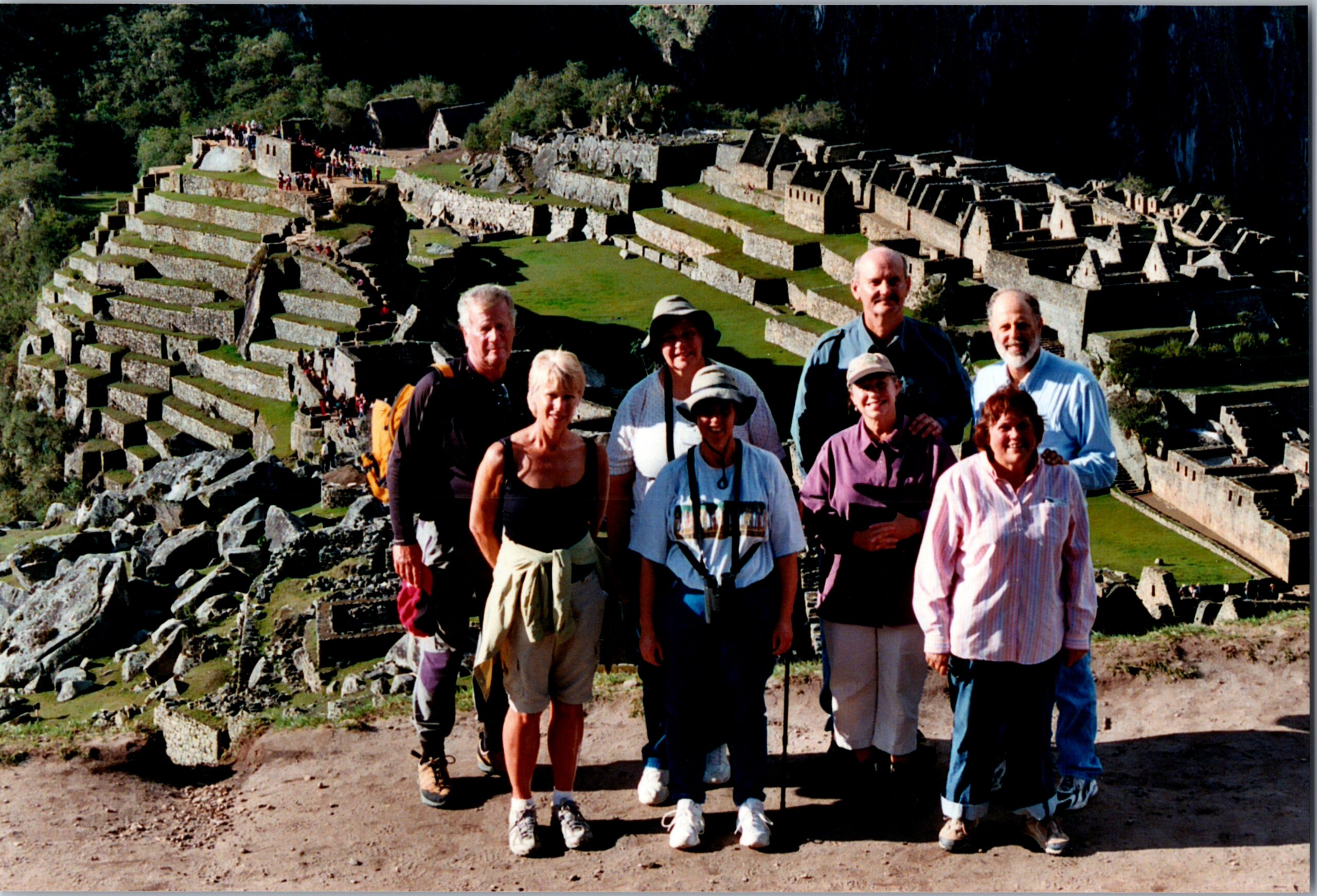 Group of smiling tourists posing together in front of ancient terraced ruins and stone buildings atop a hillside in Peru